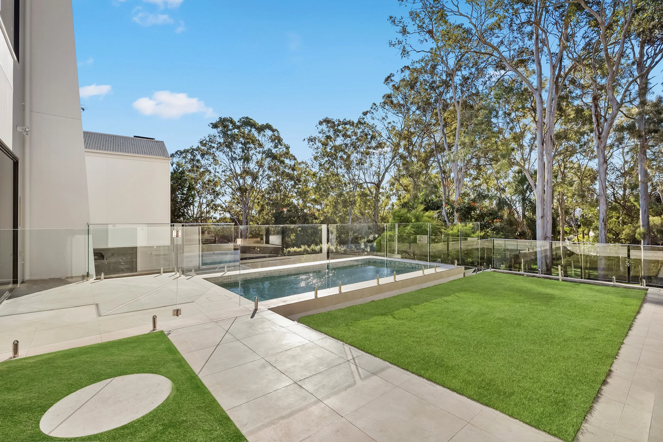 Modern backyard with swimming pool, glass fence, lush green lawn, concrete patio, and tall trees in the background under a blue sky.