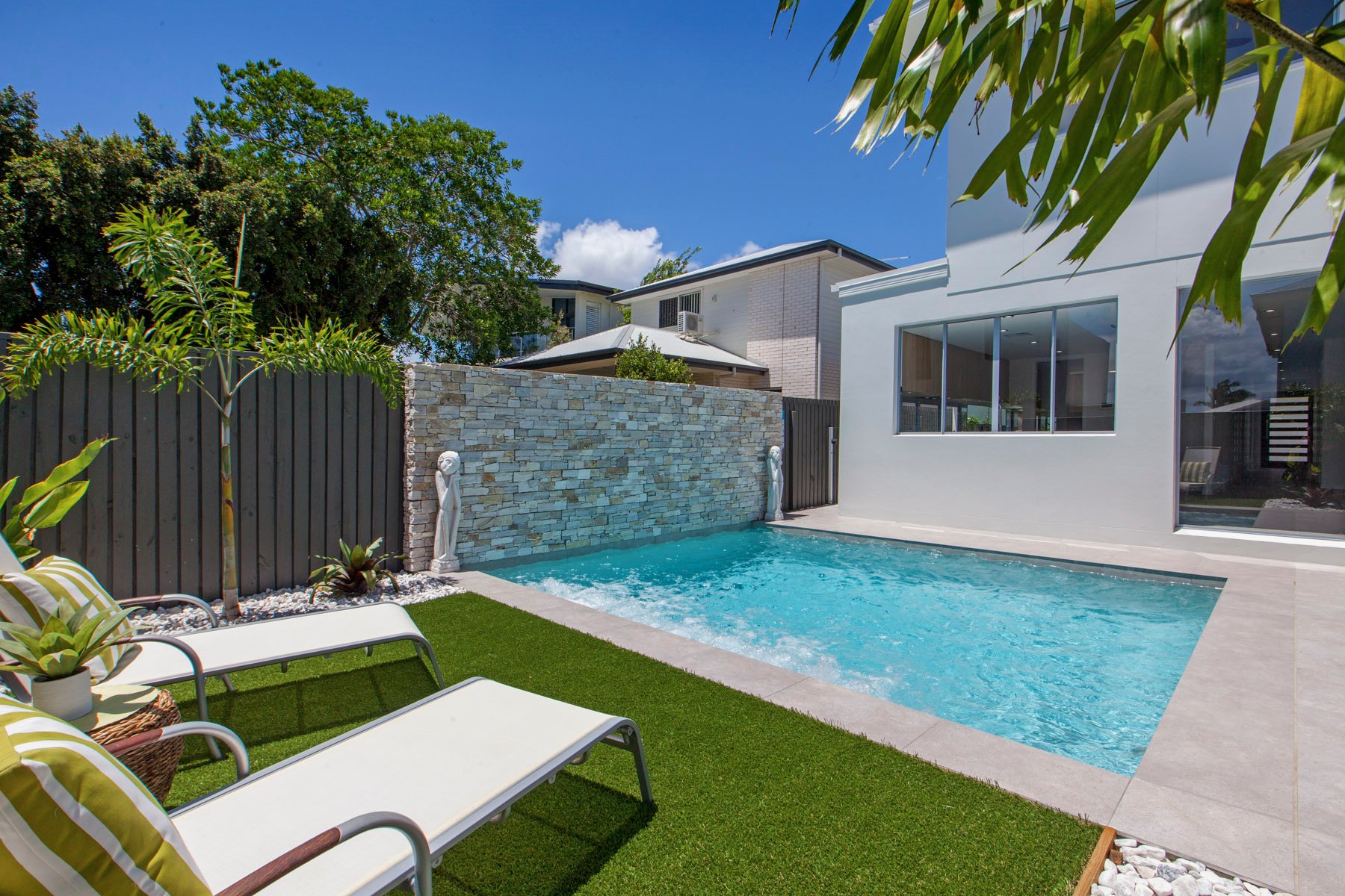Modern backyard with swimming pool, green grass, lounge chairs with striped pillows, potted plants, stone wall with sculptures, large house with glass windows, and lush trees under a blue sky.