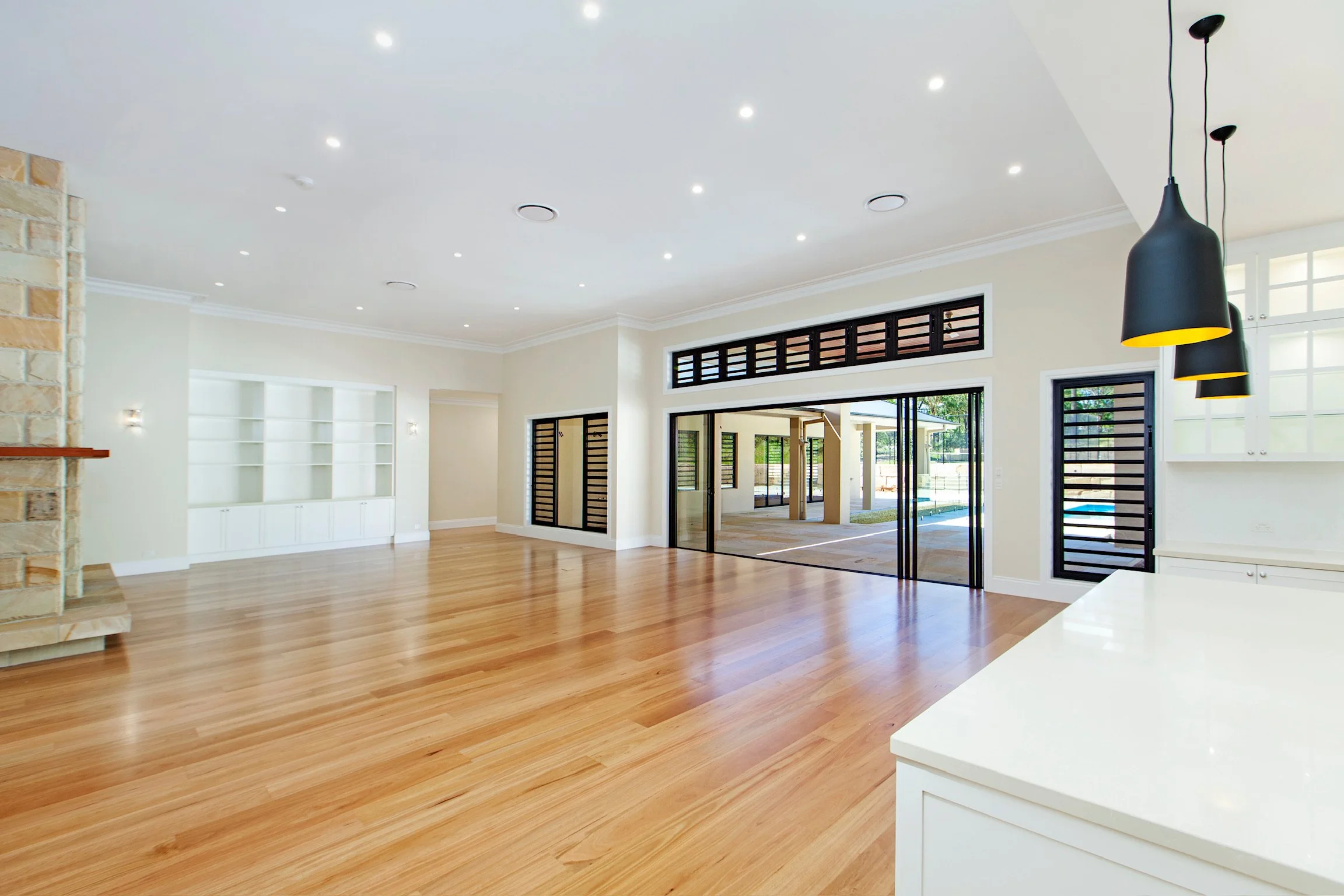 Open living room with hardwood floors, white walls, a built-in white bookshelf, large glass sliding doors, and black pendant lights hanging from the ceiling.