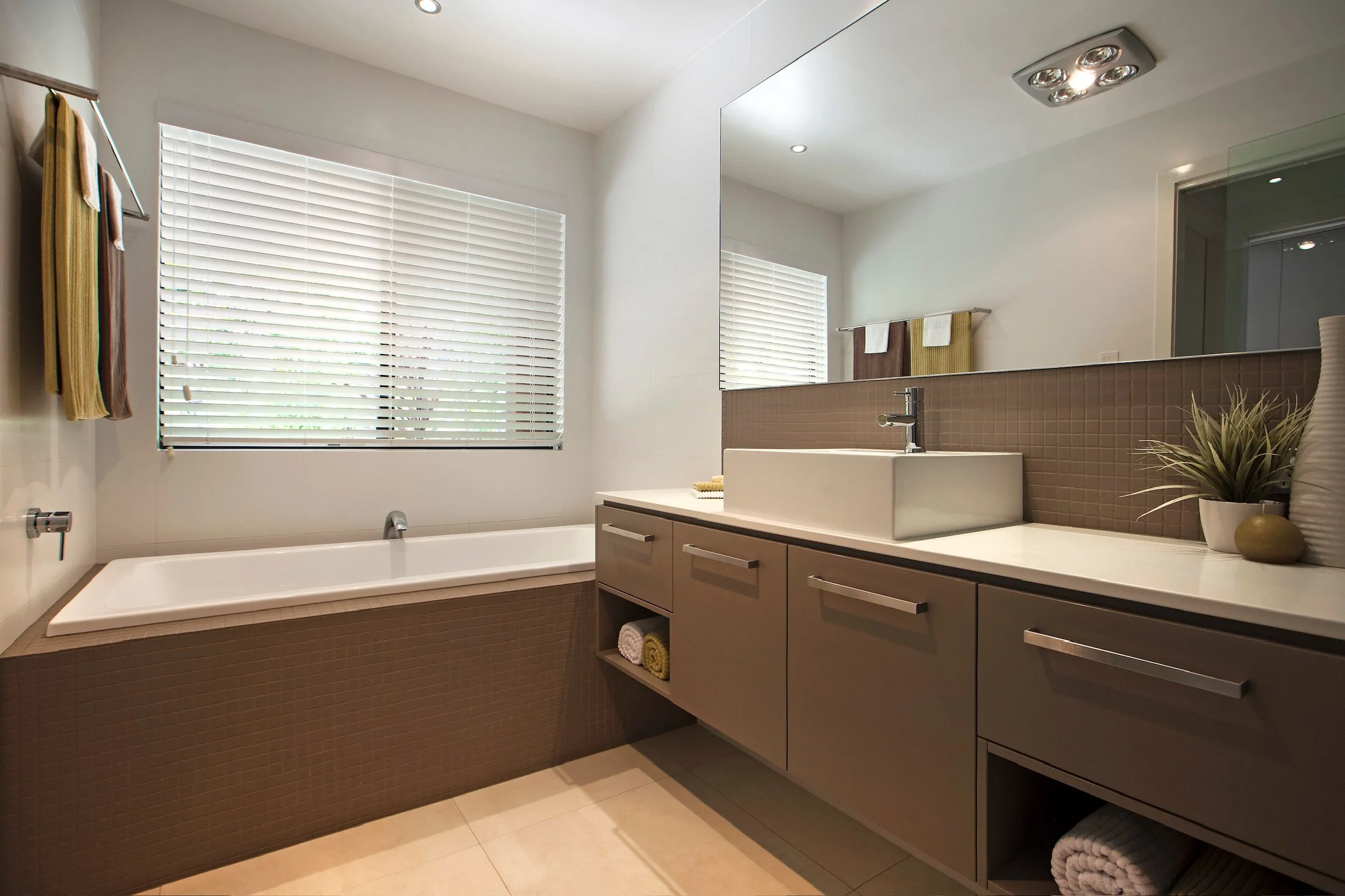 Modern bathroom with white walls, a large mirror, a white sink, a vanity with beige drawers, a bathtub, and a window with white blinds. Decor includes towels hanging on a towel bar, rolled towels in a shelf, a small plant, and a vase.