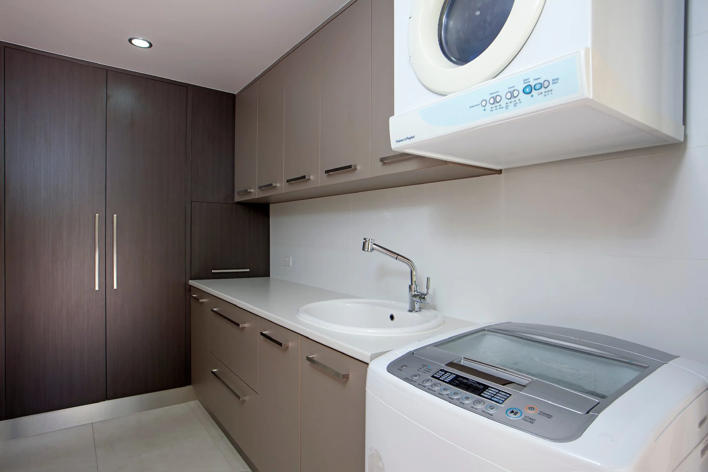 A laundry room with dark wood cabinets, a white countertop, a white sink with a chrome faucet, a washing machine, and a Fisher & Paykel dryer or washer with control panel on top.