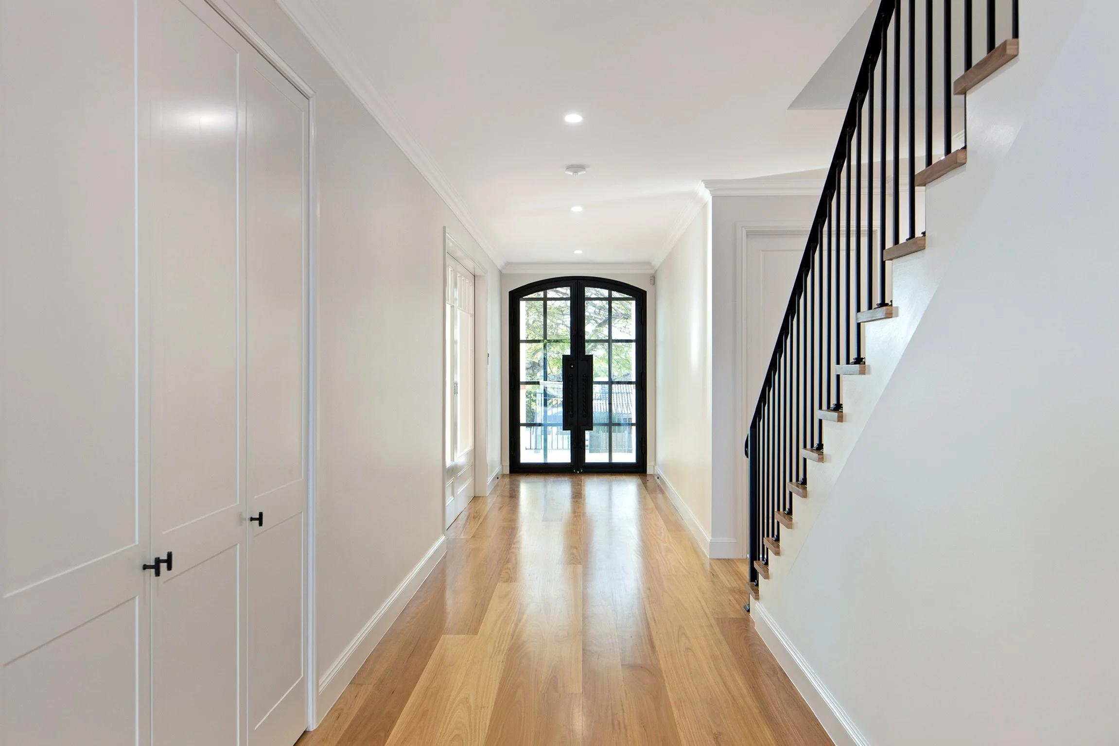 Bright hallway with hardwood flooring, white walls, built-in cabinets on the left, and a staircase with black balusters and wooden handrails on the right. A black front door with large glass panes is at the end of the hallway, with windows on either 