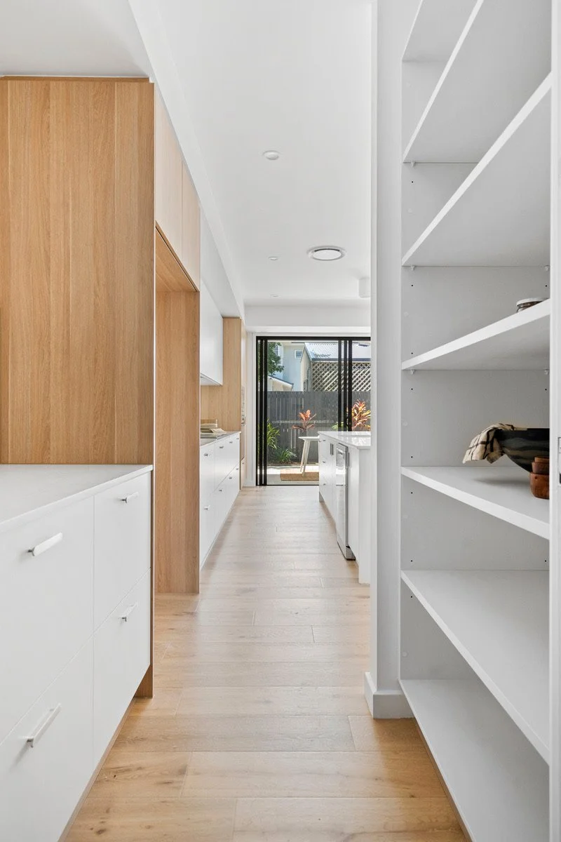 Interior view of a modern kitchen with white cabinets, wooden paneling, and open shelves, leading to a sliding glass door with a small outdoor deck and greenery outside.
