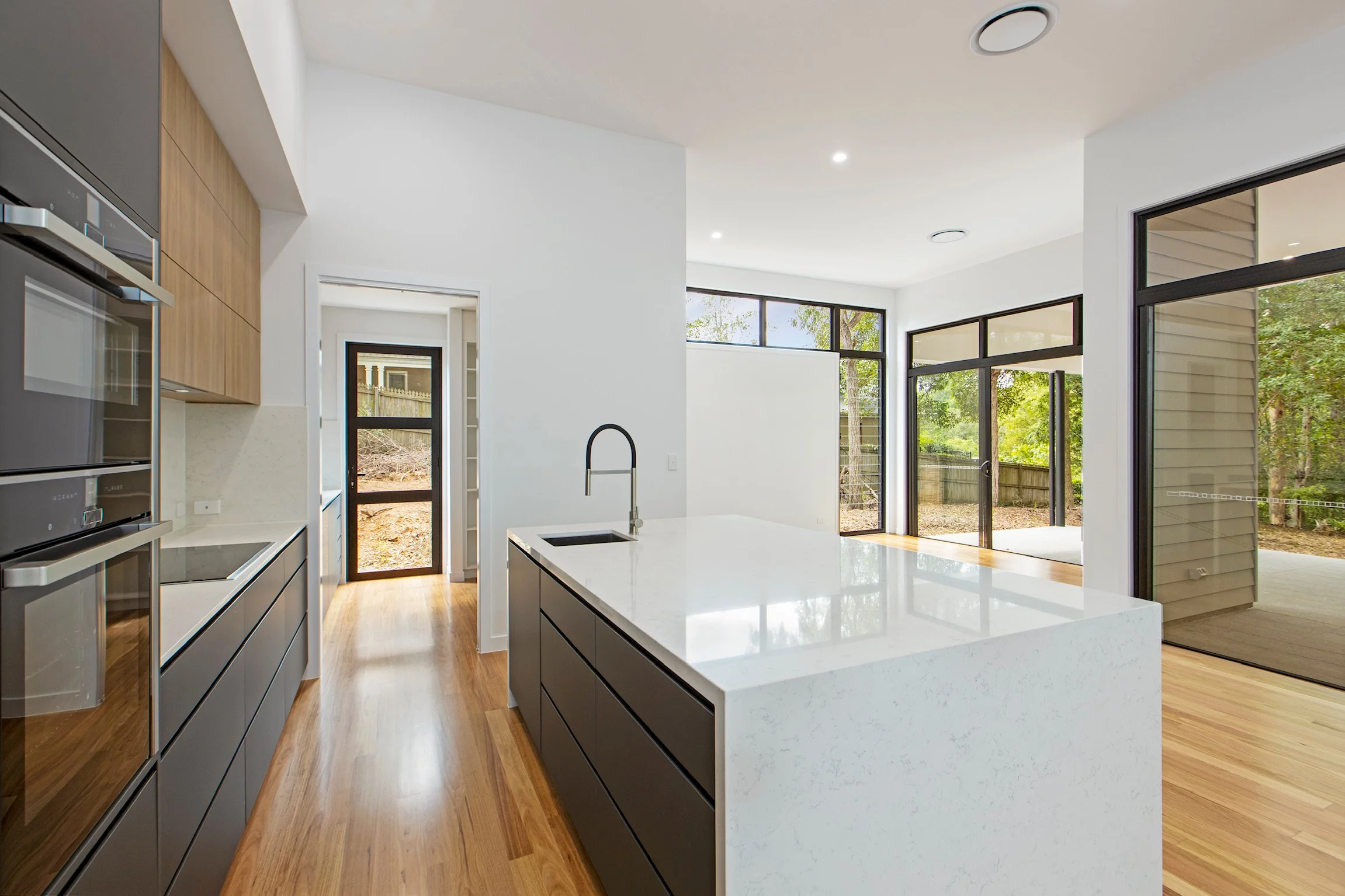 Modern kitchen with white countertops, black lower cabinets, wooden upper cabinets, and large windows providing natural light and views of a backyard.