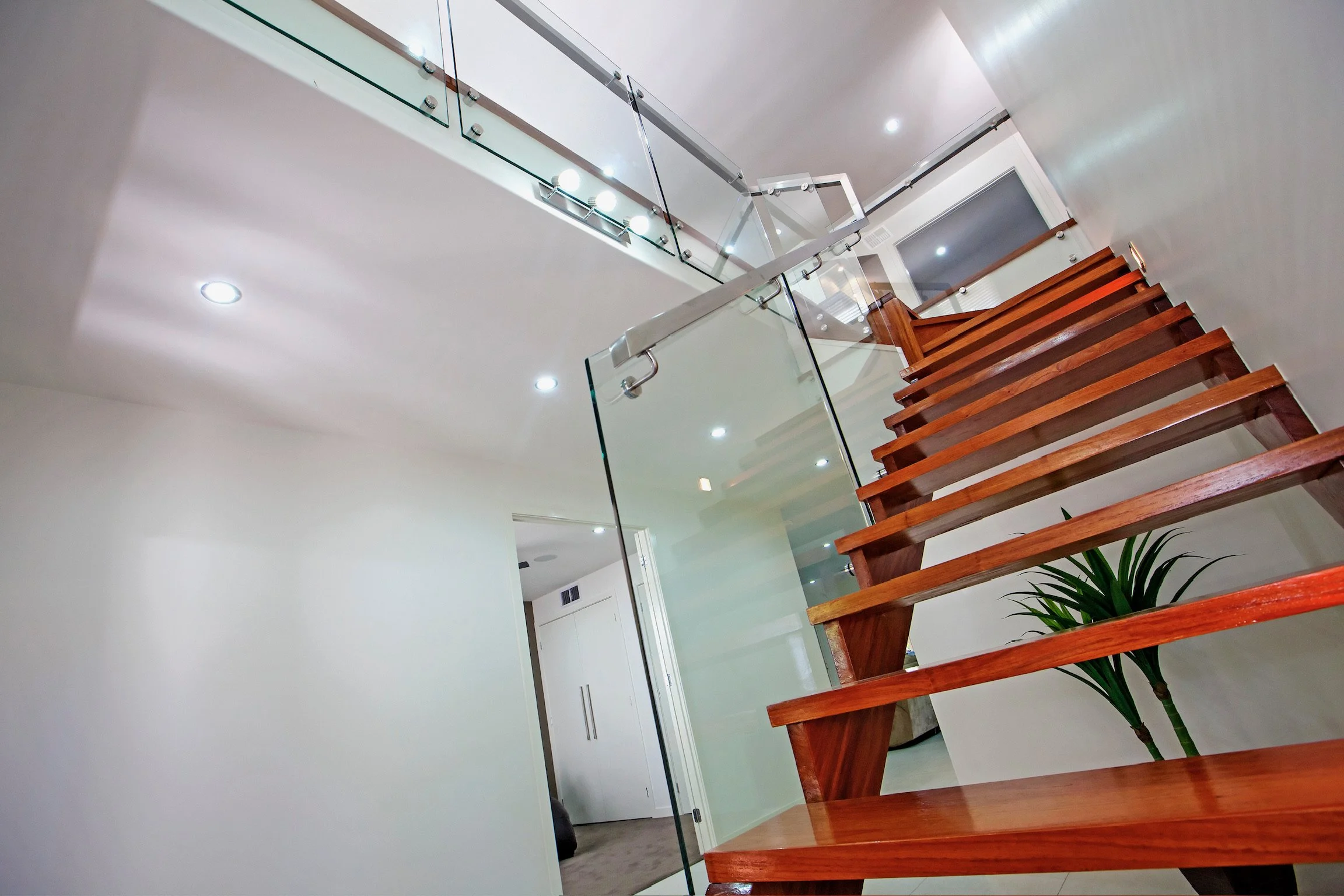 Interior view of a modern staircase with wooden steps, glass railing, and ceiling lights, showing part of a white wall and door in the background.