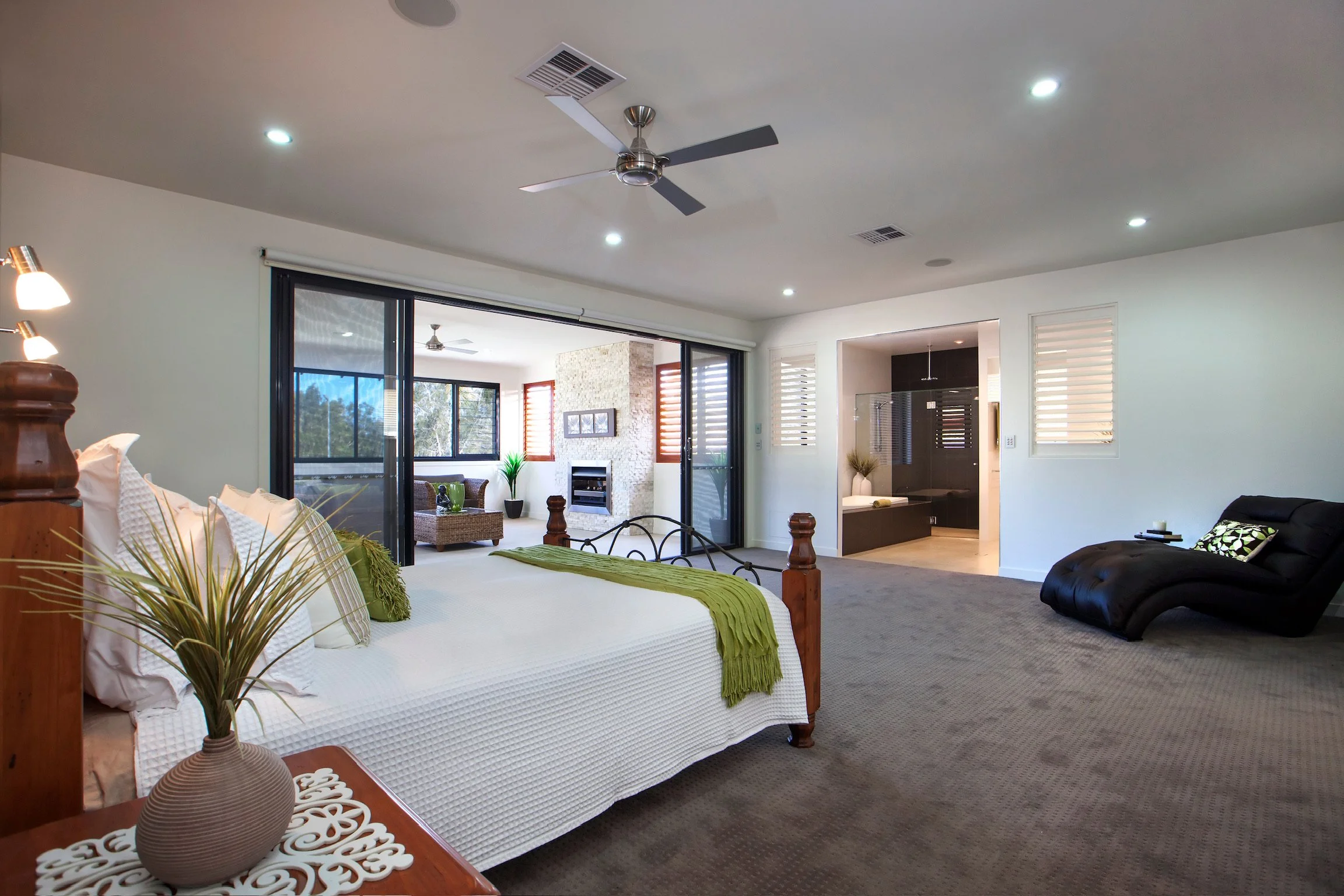 A spacious bedroom with a white bed featuring pillows and a green throw blanket, a bedside table with a decorative vase, and a view into a sitting area with a fireplace and large windows.