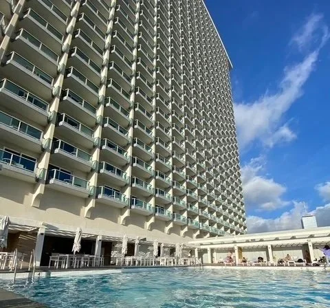 View of a large hotel or apartment building with a swimming pool in the foreground on a sunny day.