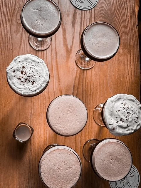 Top-down view of six beer glasses with foam on top and two whipped cream desserts with chocolate sprinkles on a wooden table.