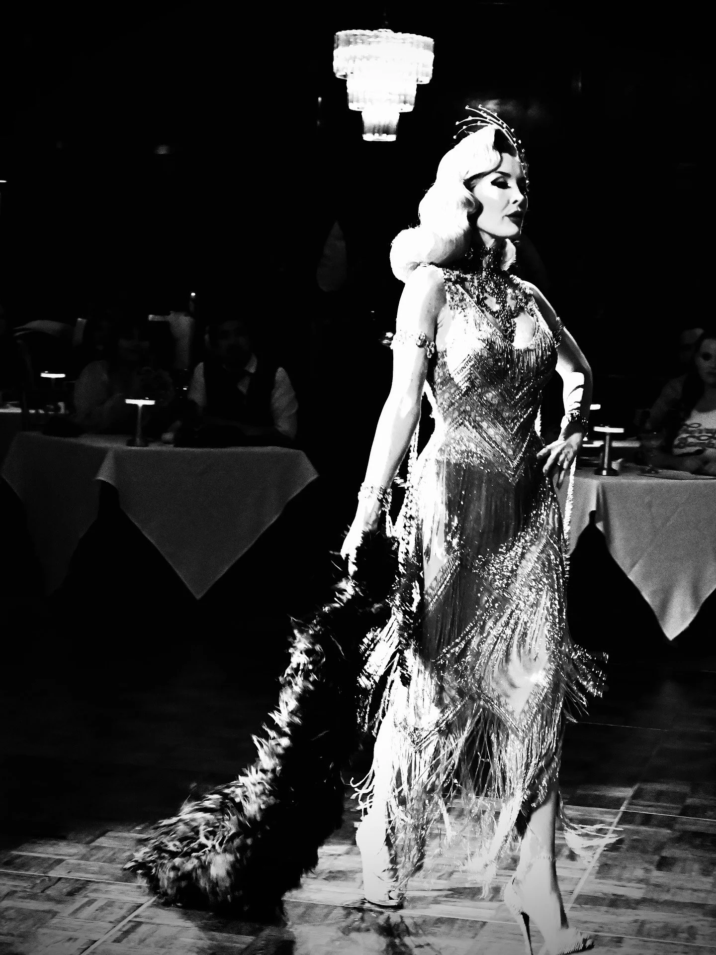 A woman in an elegant, shimmering gown with intricate beadwork and feathers, walking across a stage or dance floor. She has styled hair, bold makeup, and is wearing jewelry, holding a fur or feathered accessory in her hand. The background appears to be a dimly lit room with people seated at tables, possibly in a performance or event setting.