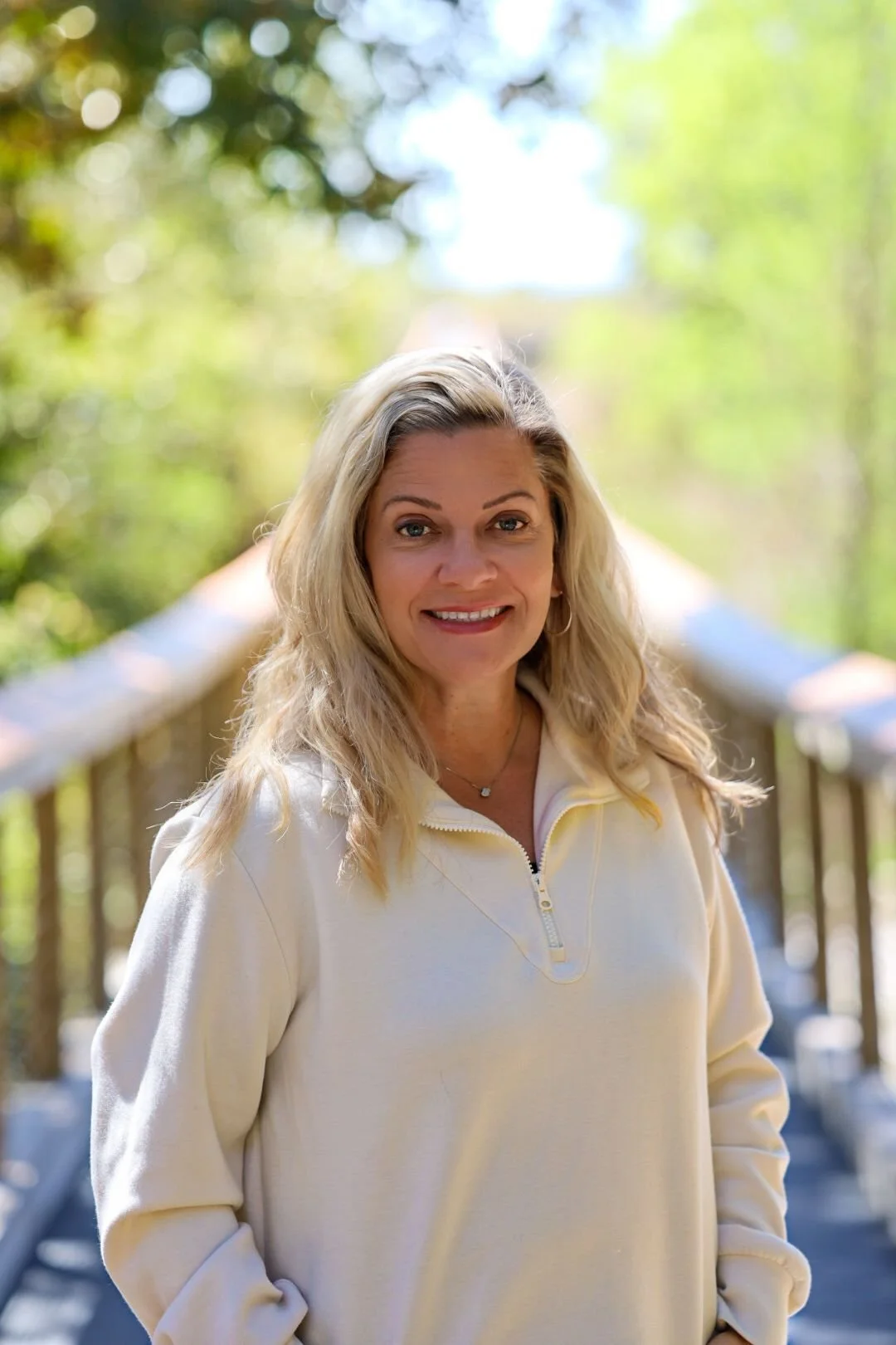 A smiling woman with blonde hair standing outdoors on a wooden bridge surrounded by green trees and bright sunlight.