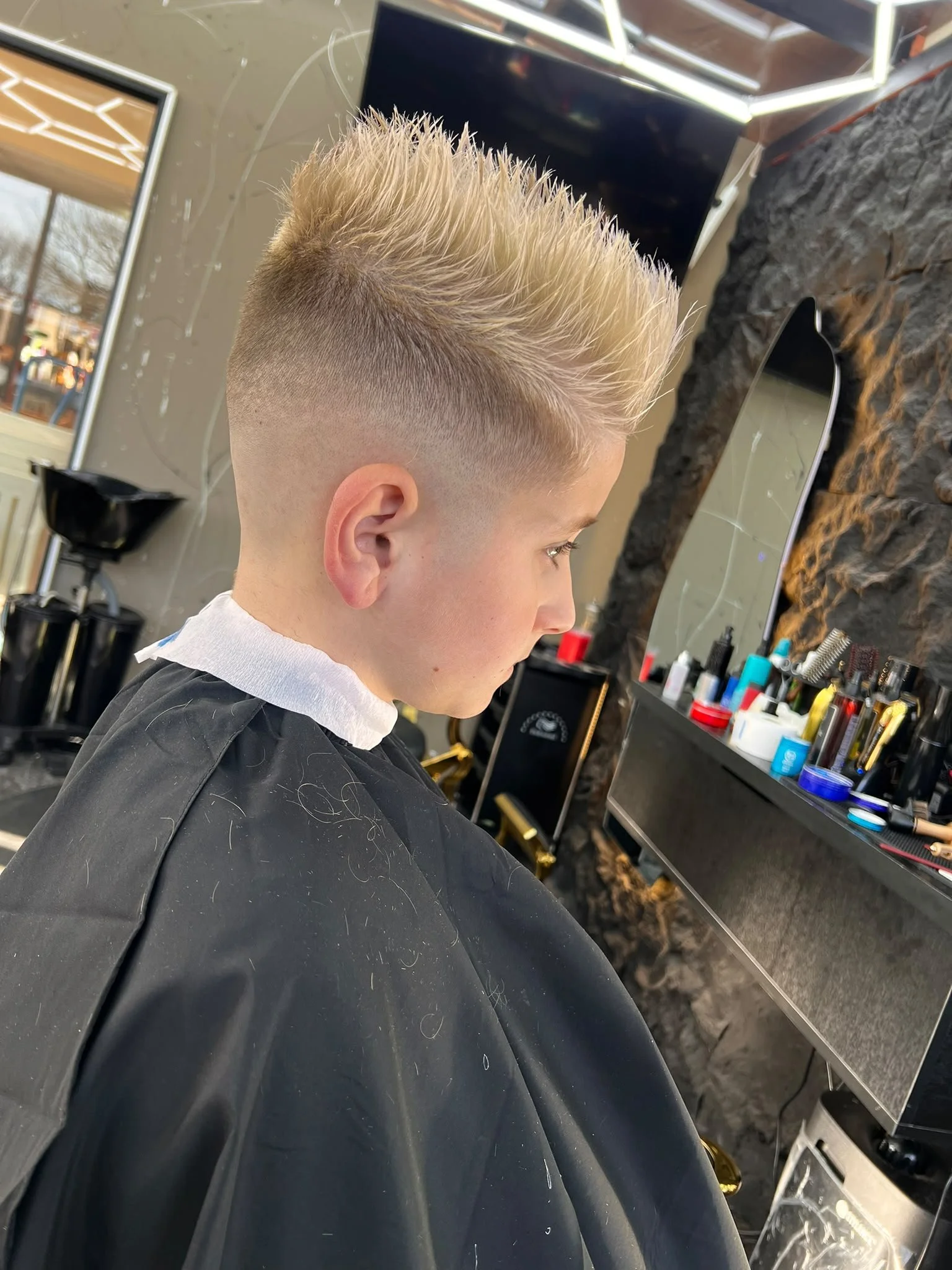 Side view of a young man with bleached blonde spiked hairstyle getting a haircut in a barbershop, with shelves of hair products and a mirror in the background.