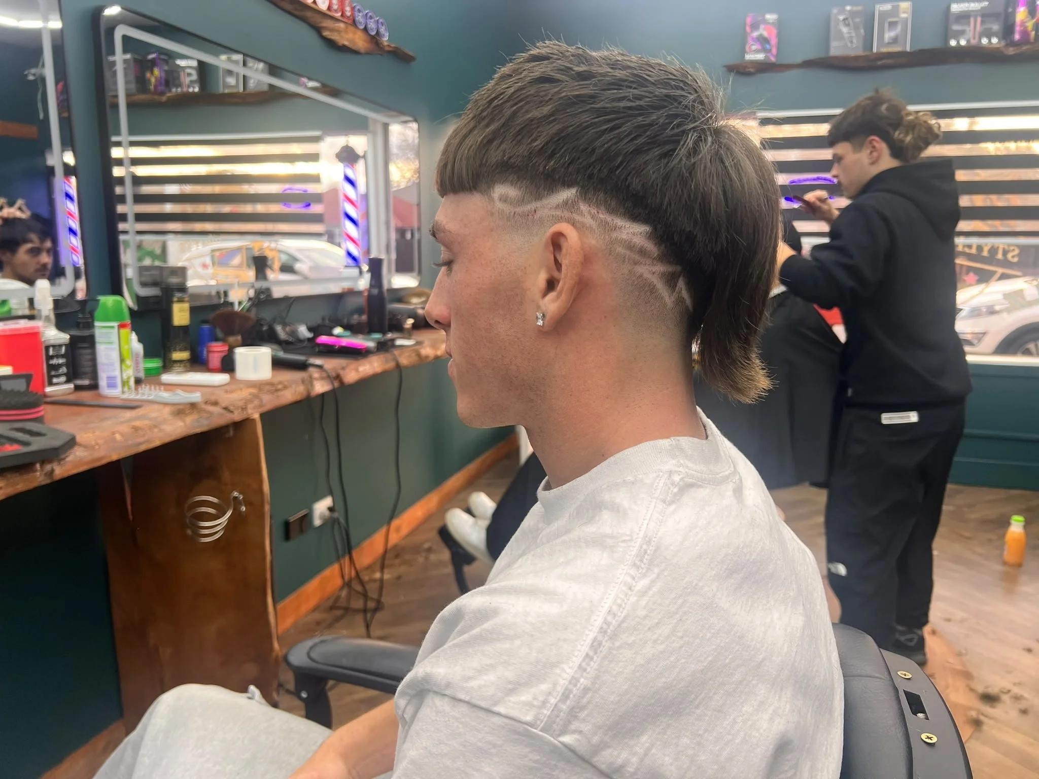 A person with a short, textured haircut with a shaved design on the side, sitting in a barber's chair at a barbershop. The barber is in the background, working on another client. The barbershop has a mirror, shelves with hair products, and a wooden counter with grooming tools.