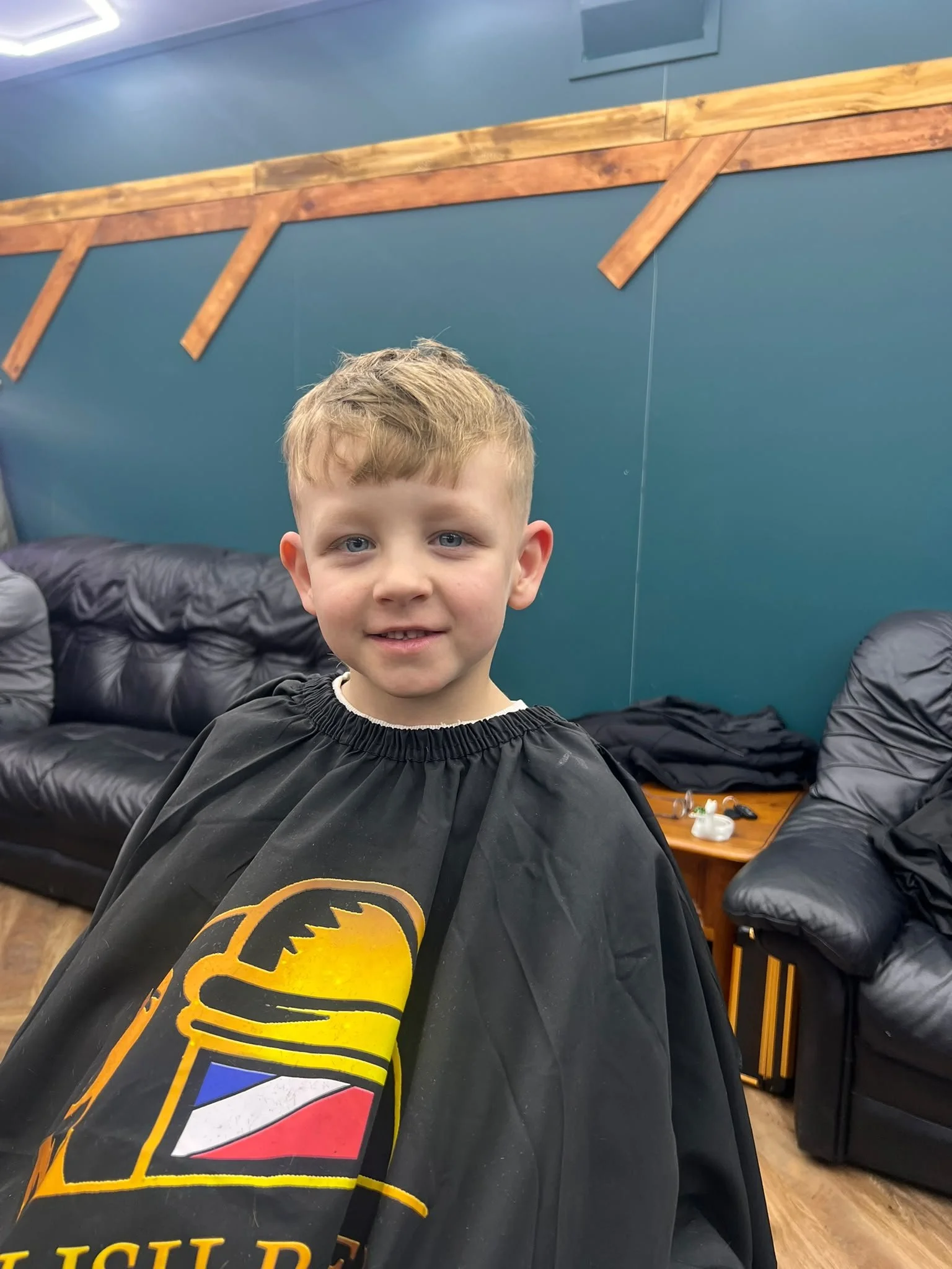 A young boy with light brown hair and blue eyes sitting in a salon chair, wearing a black cape with a Batman logo, in a room with blue walls and black leather couches.