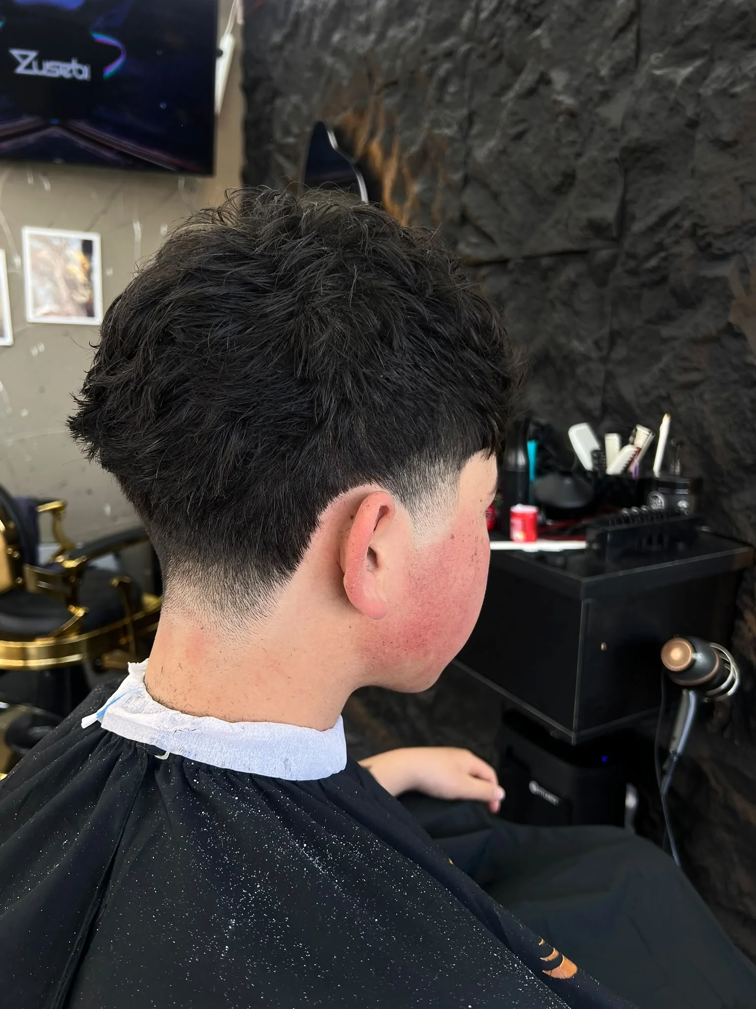 A man with dark, curly hair sitting in a hair salon chair, preparing for a haircut or styling session, with salon equipment and a textured dark wall in the background.