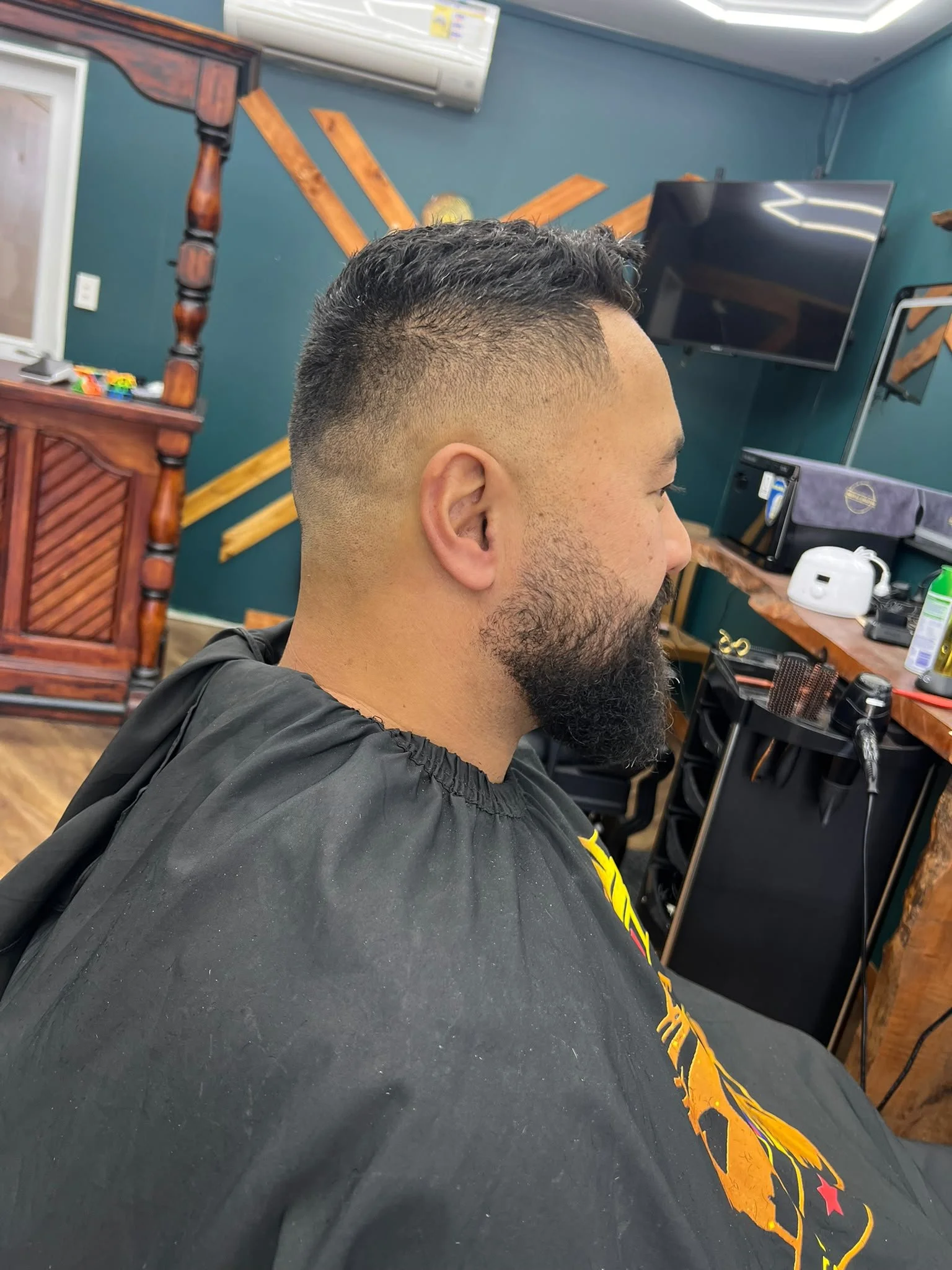 Side profile of a man with a freshly cut fade hairstyle and a beard sitting in a barbershop. The background features a television, a wooden shelf with various items, and a teal wall with wooden accents.