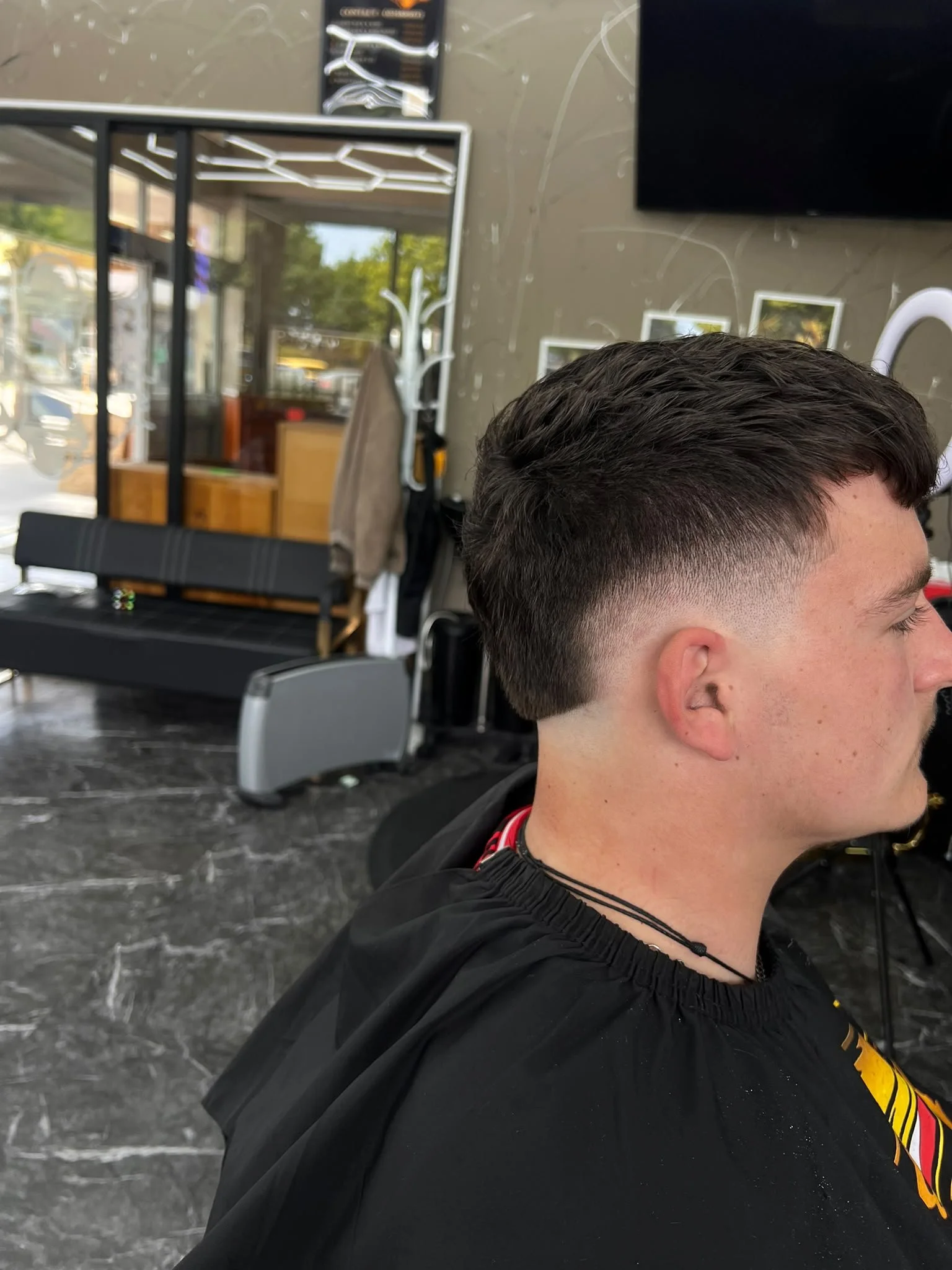 The image shows a young man with short, dark styled hair, sitting in a barbershop or hair salon with a black cape around his shoulders. In the background, there are salon chairs, a mirror, and various framed pictures on the wall. The setting has a modern, professional appearance with large windows letting in natural light.