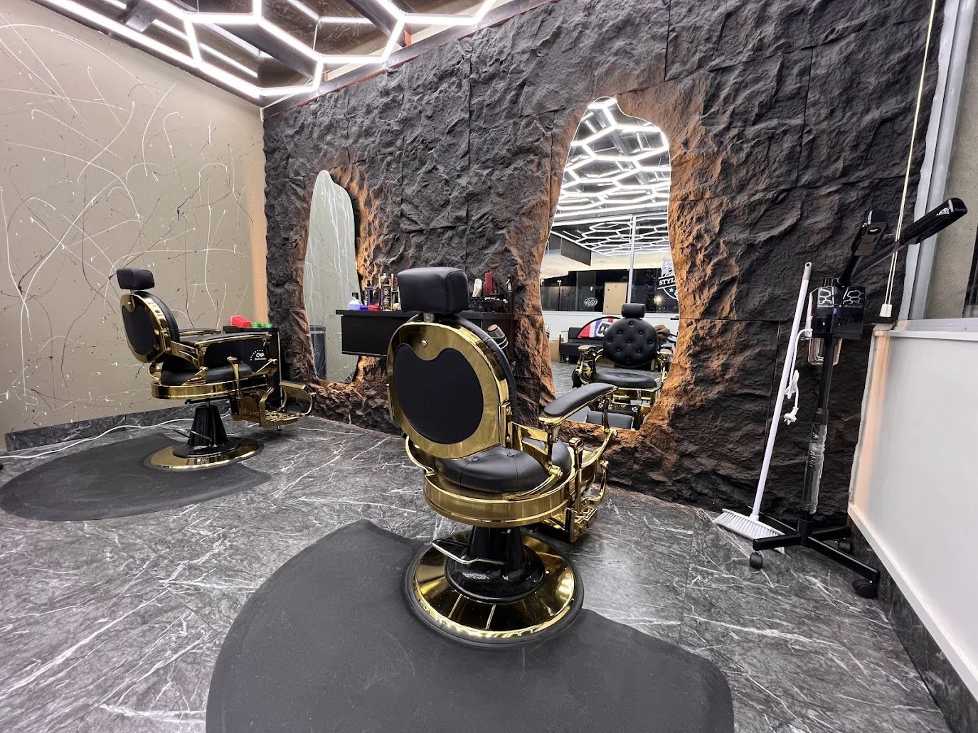 Inside a modern barbershop with two vintage-style barber chairs with black leather seats and gold accents, a textured black rock wall with two mirrors, and bright LED ceiling lights.
