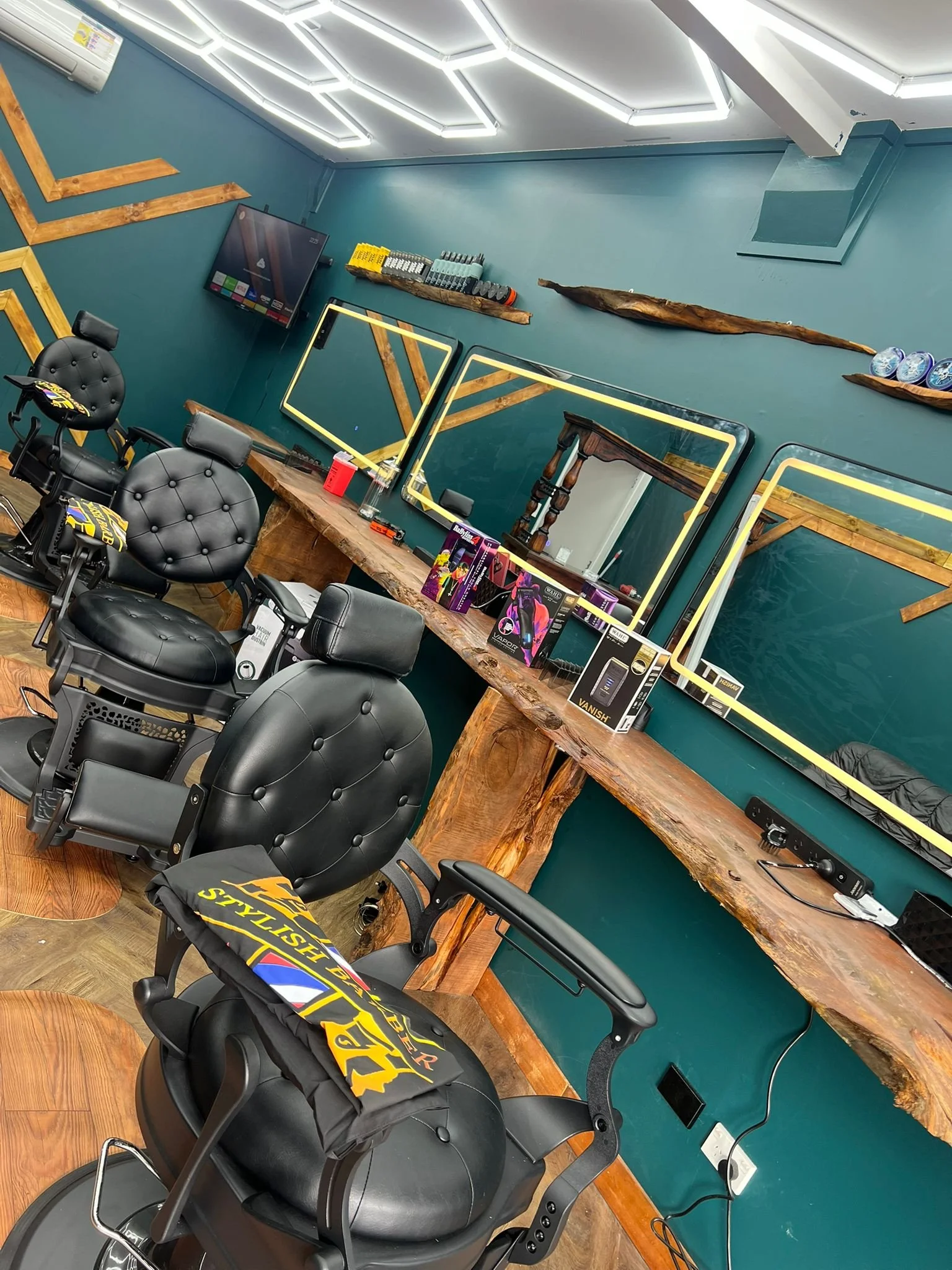 Barbershop or salon workspace featuring black barber chairs, large wooden worktables, mirrors with black and yellow frames, and a teal wall with decorative wooden accents and a mounted TV.