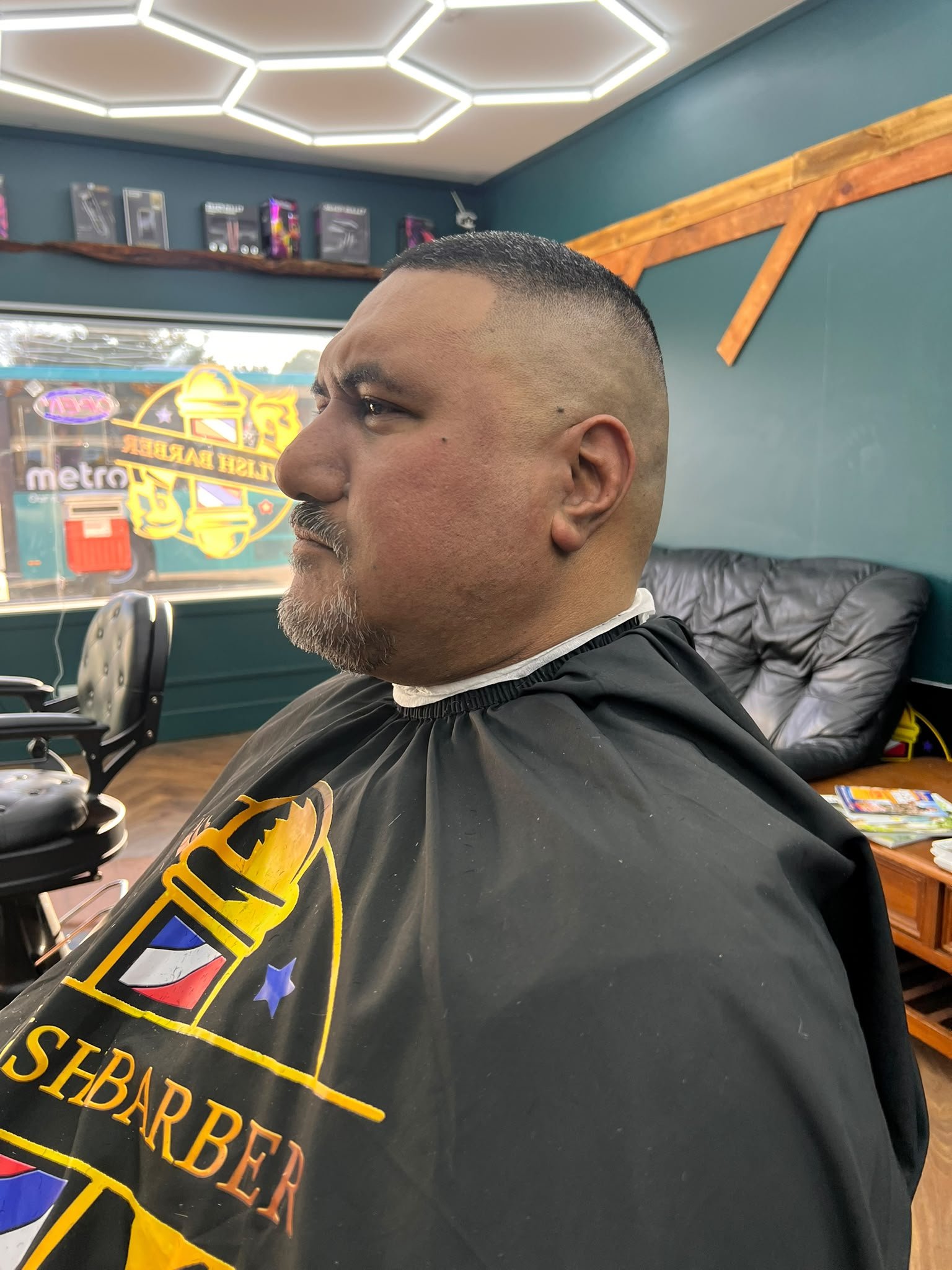 Man with a beard and short hair seated in barber shop, wearing a barber cape with a logo, in front of large window with colorful amusement park poster.