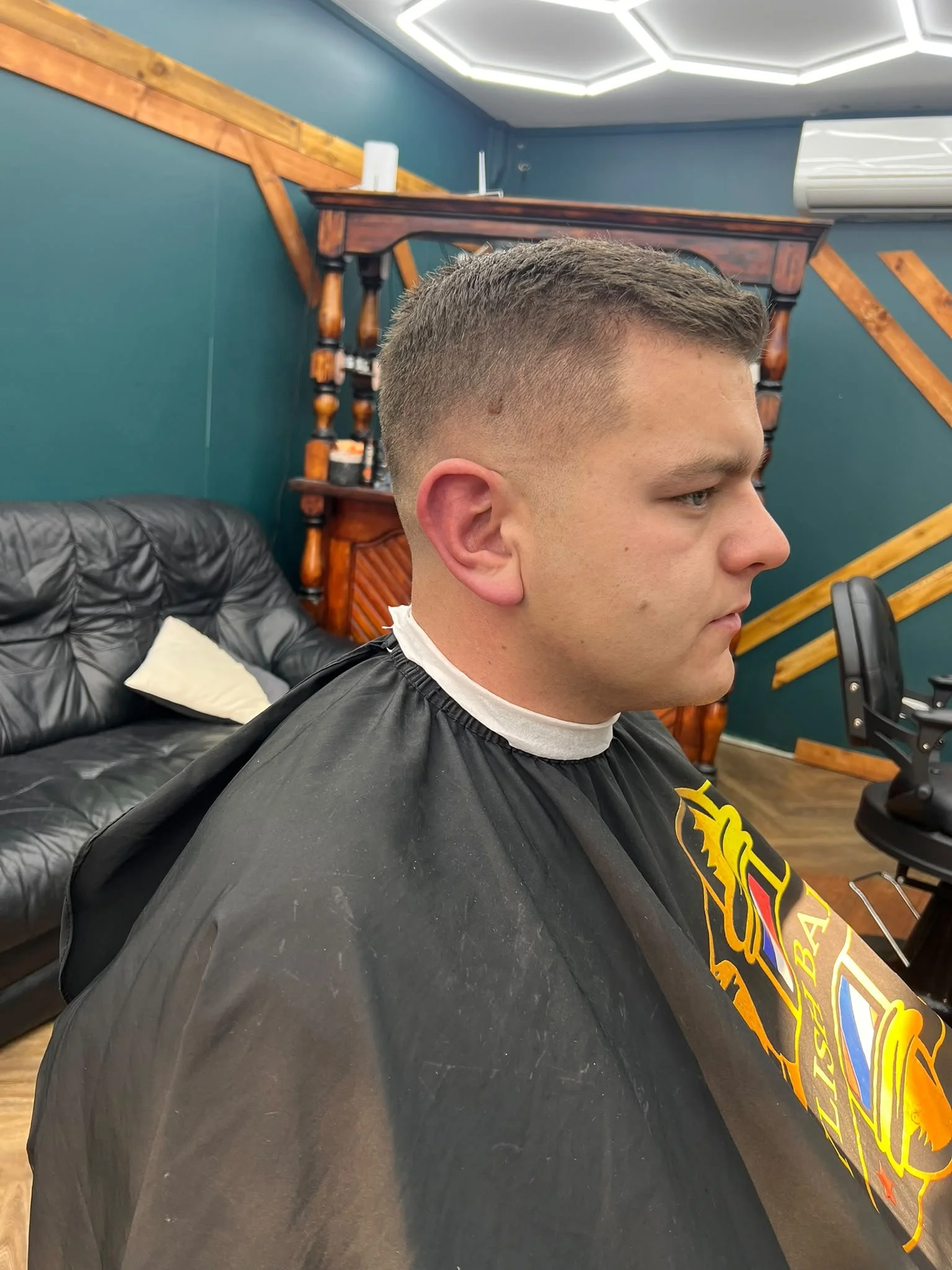 A young man getting a haircut at a barbershop, sitting in a black barber cape with a tattoo on his right arm, against a background of blue walls, a wooden shelf and a black leather couch in a modern interior.