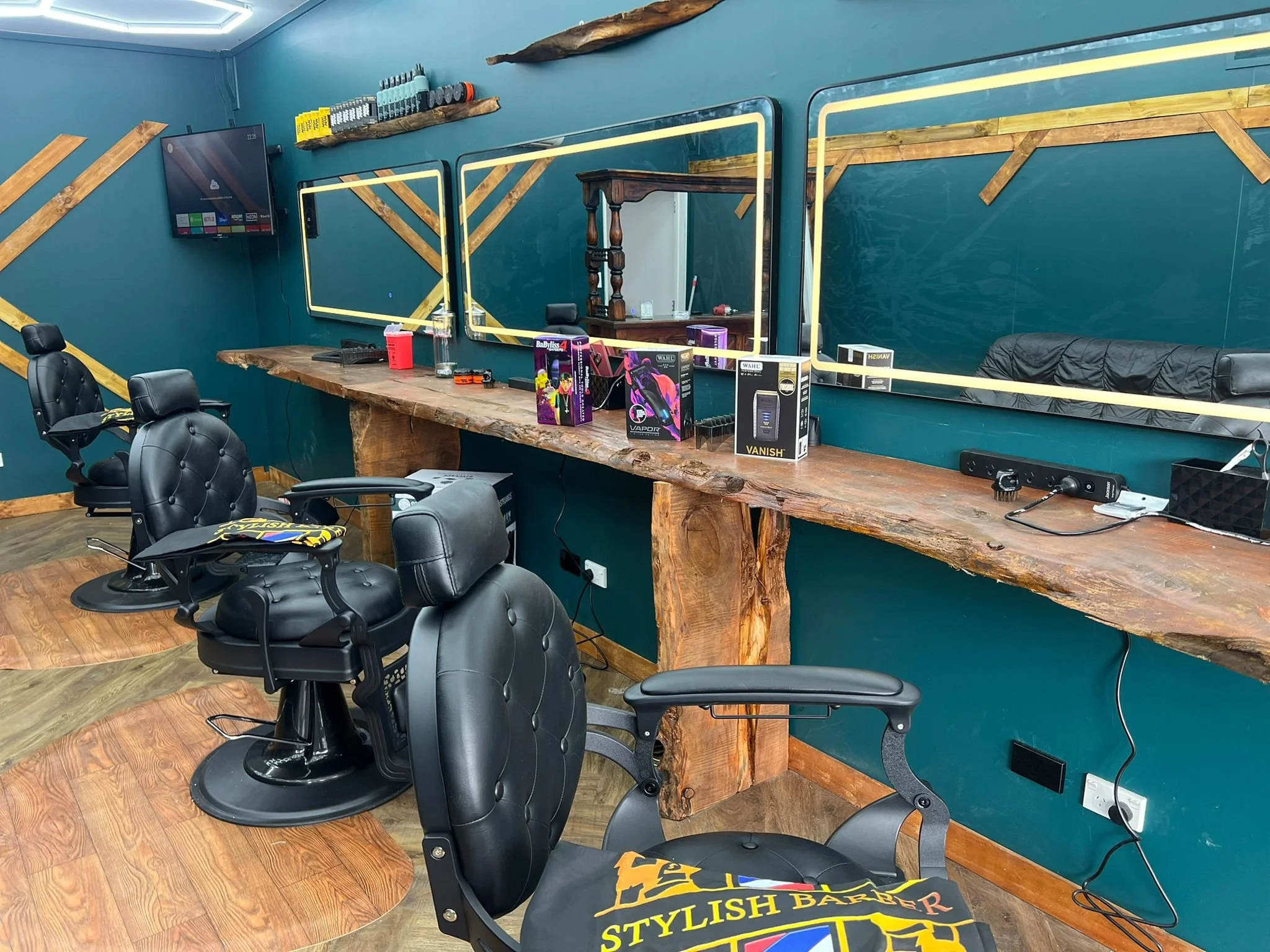A barber shop or salon with a long rustic wood counter, black barber chairs, and large mirrors framed with yellow light. The decor includes teal walls with diagonal wooden accents, a mounted TV, and shelves with hair products. Various barber tools and products are placed on the counter.