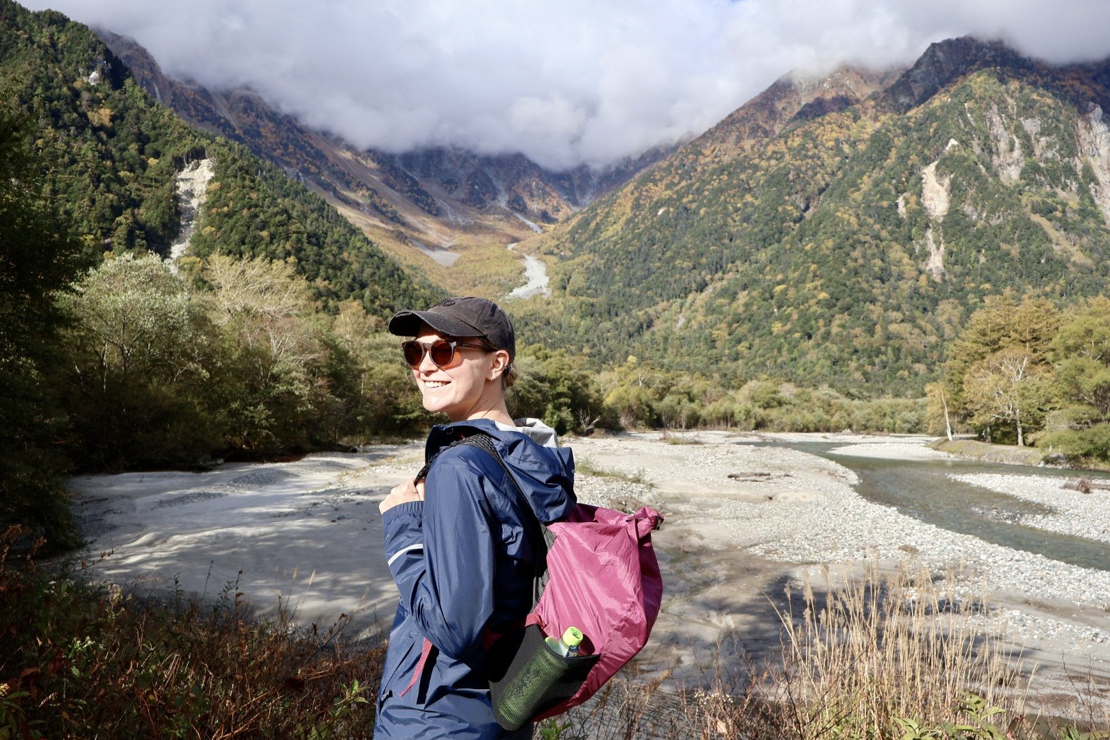 A woman outdoors in a mountainous landscape with a river and lush greenery, smiling, wearing sunglasses, a black cap, and carrying a pink backpack.