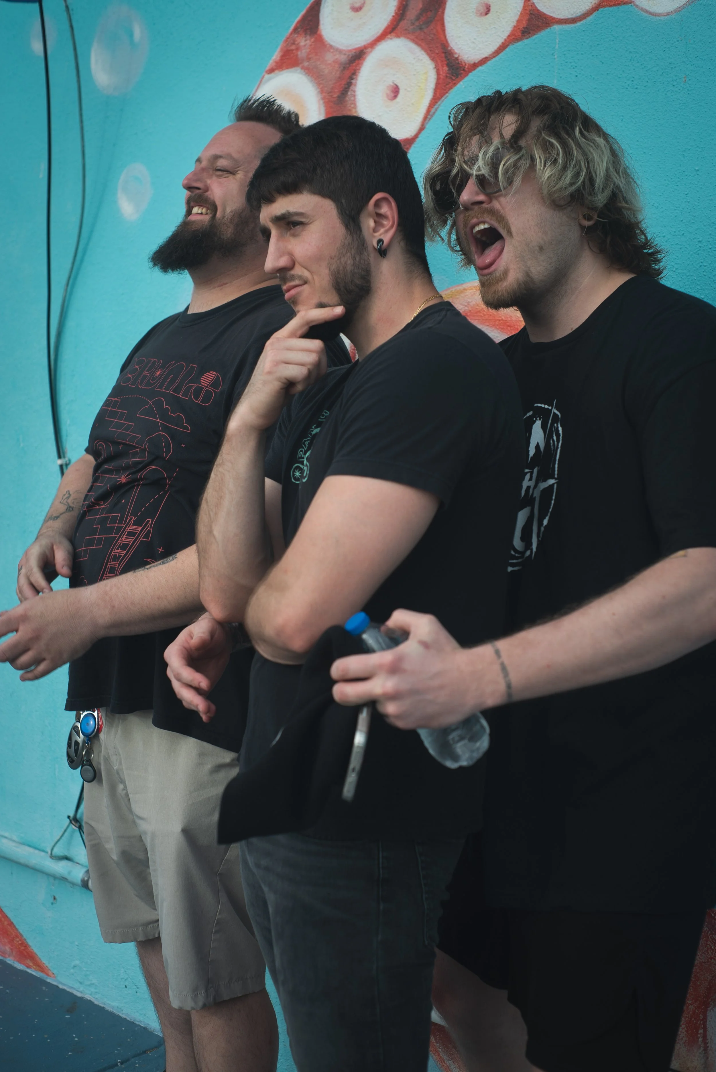 Three young men standing against a blue wall with colorful artwork. The man on the left has a beard and is smiling with closed eyes, wearing a black T-shirt. The man in the middle has dark short hair, a beard, and is in a thoughtful pose with his hand on his chin, wearing a black T-shirt. The man on the right has curly blond hair, is wearing sunglasses and a black T-shirt, and is shouting or singing with his mouth open.