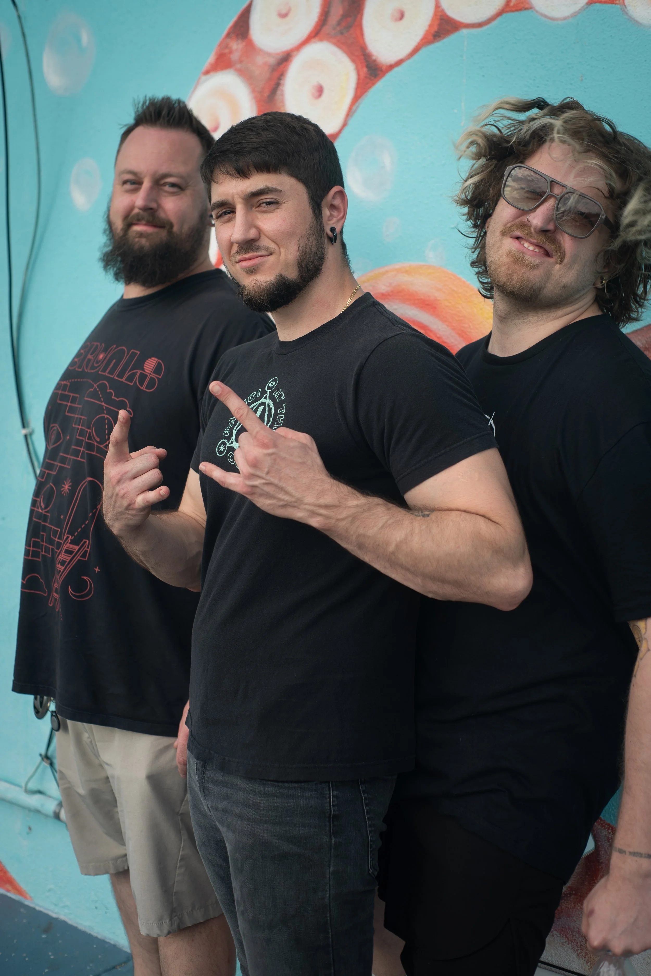 Three men standing in front of a colorful mural, with the man in the center making a hand gesture, all three looking towards the camera.