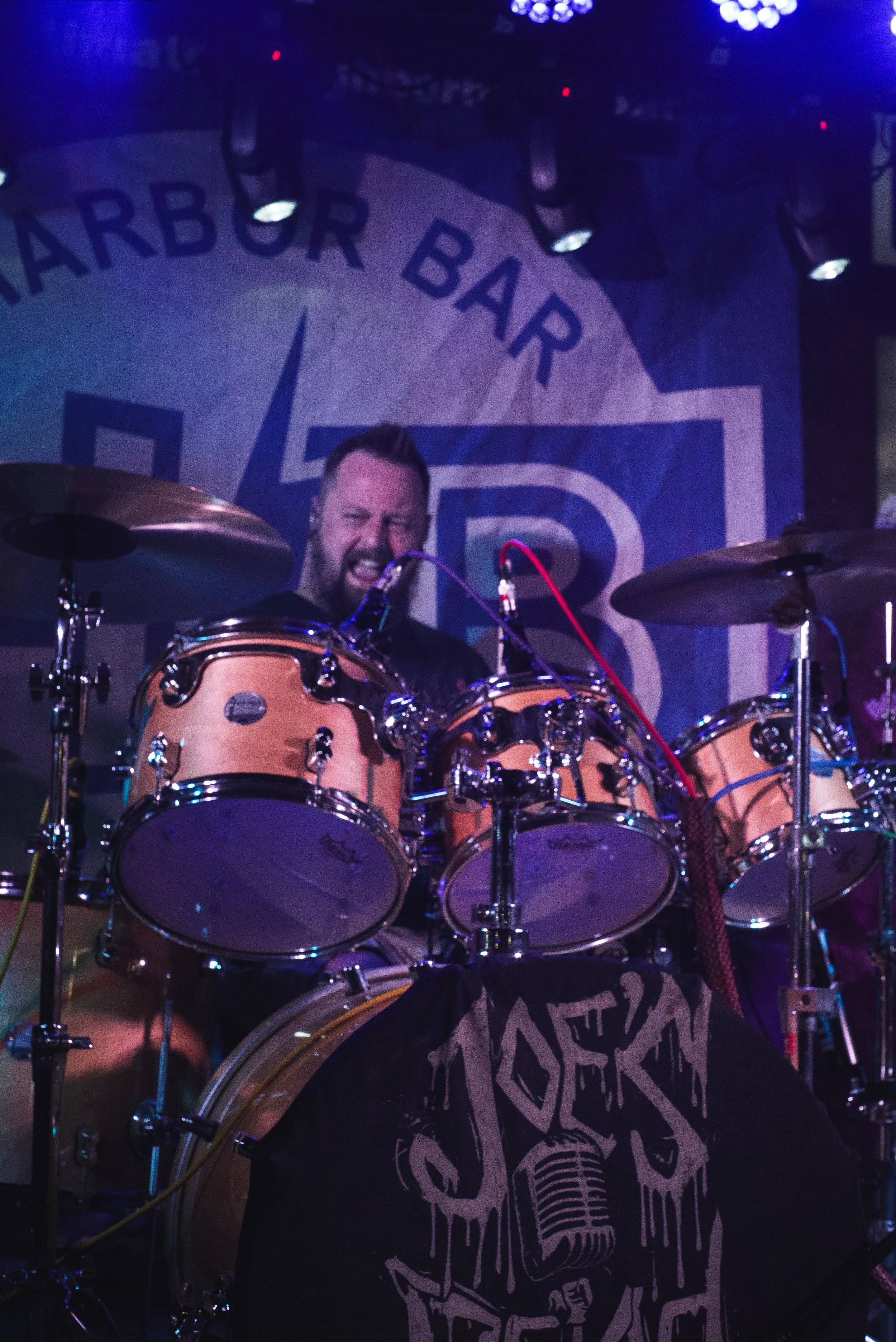 Male drummer playing on stage with drums and cymbals, with a background sign that reads "Harbor Bar." The stage is lit with blue and purple lights.