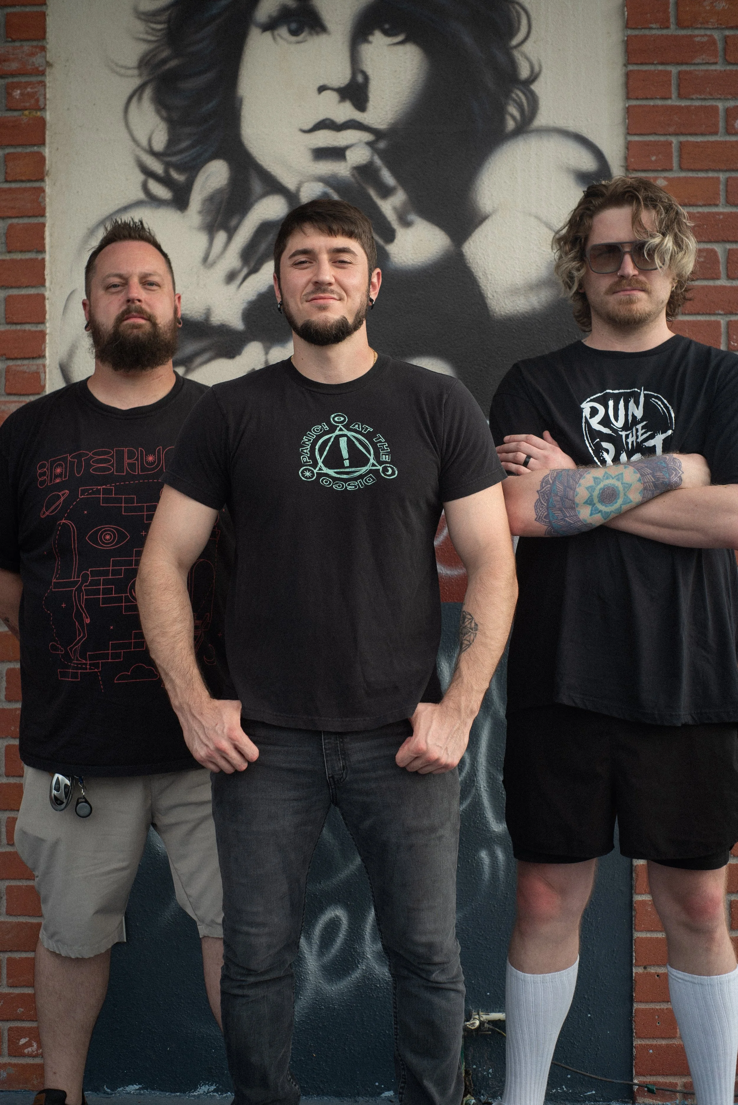 Three young men standing in front of a large black and white mural of a woman with curly hair, one wearing headphones around his neck, another with sunglasses and tattoos, and the third with a black t-shirt with graphics.