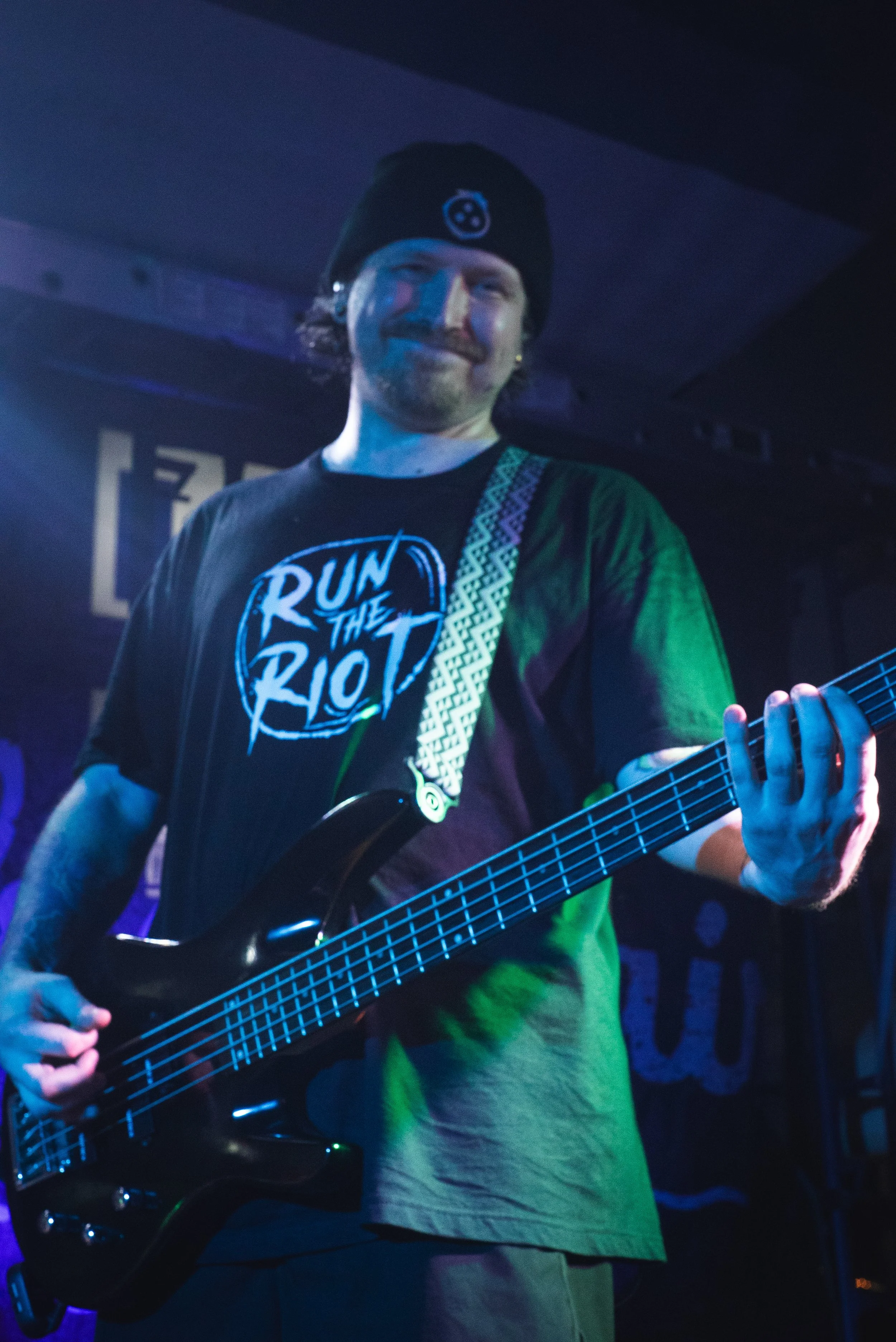 A man smiling while playing an electric bass guitar on stage. He is wearing a black beanie, a black t-shirt with white text, and dark pants, with stage lighting creating a bluish and purple ambiance.