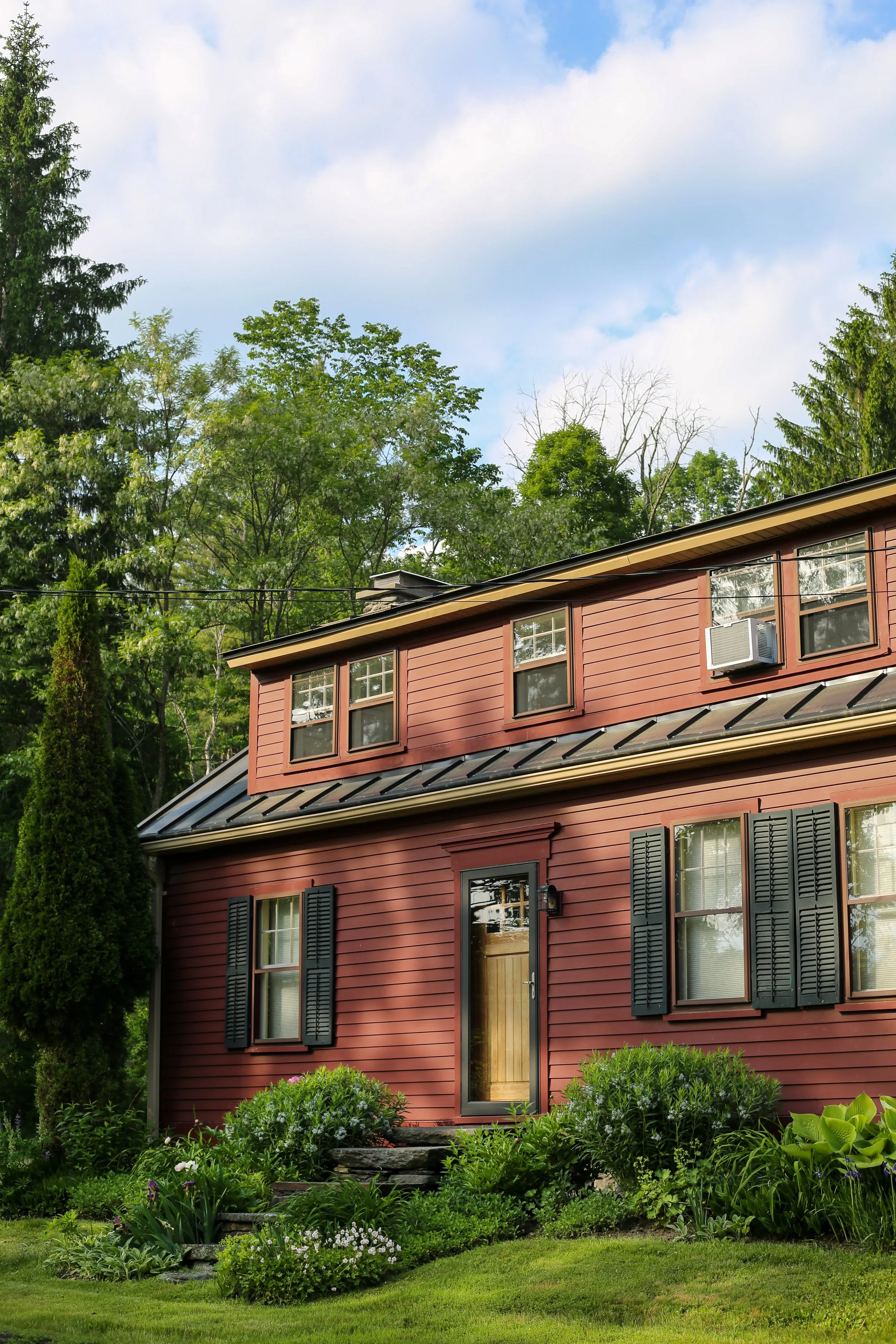 A red two-story house with black shutters and a wooden front door, surrounded by a well-maintained garden with various green plants, bushes, and flowers. Tall trees and a partly cloudy sky are visible in the background.