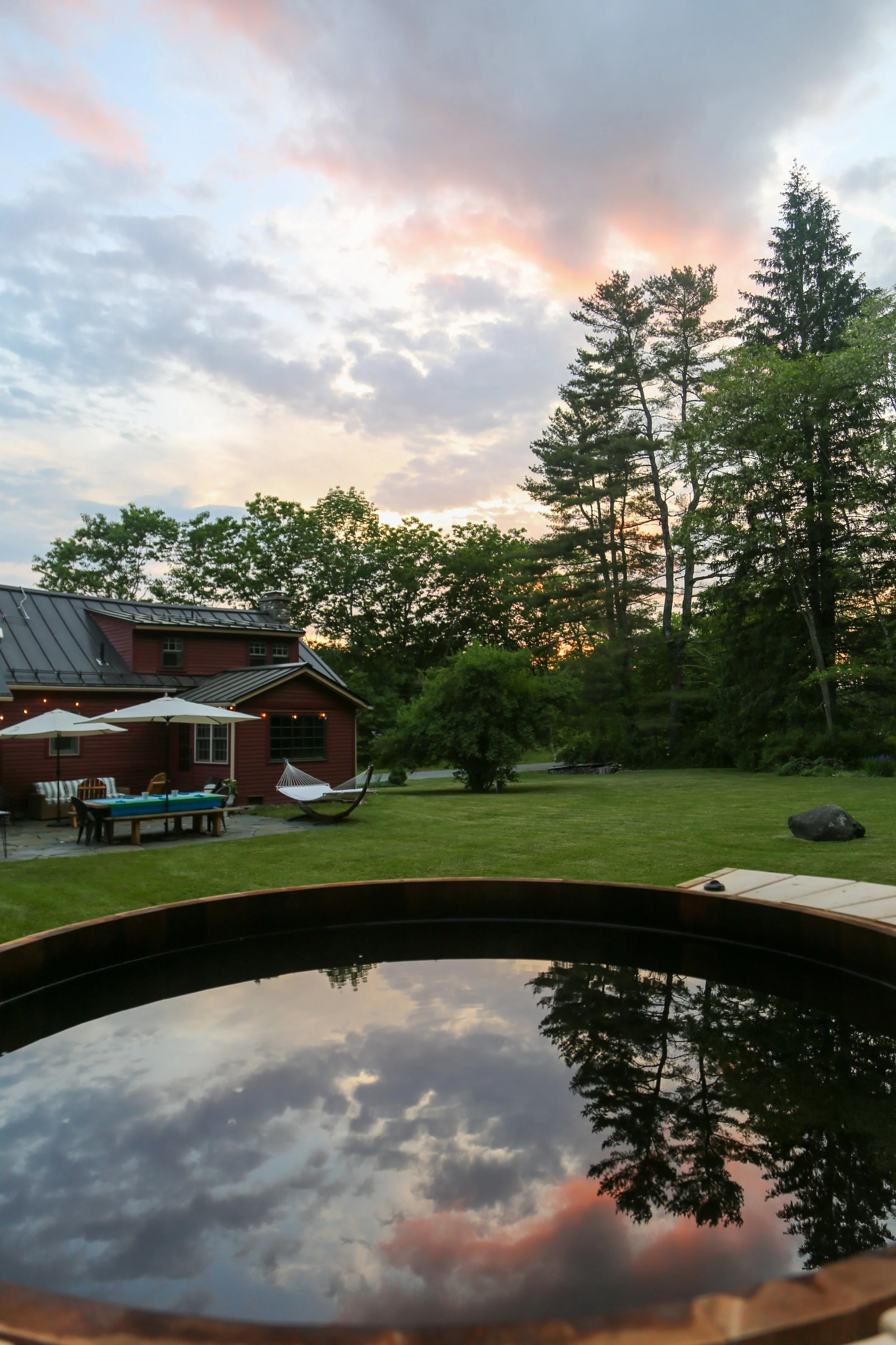 A view of a backyard at sunset with tall trees, a red house, patio furniture, and a reflective surface in the foreground reflecting the sky and clouds.