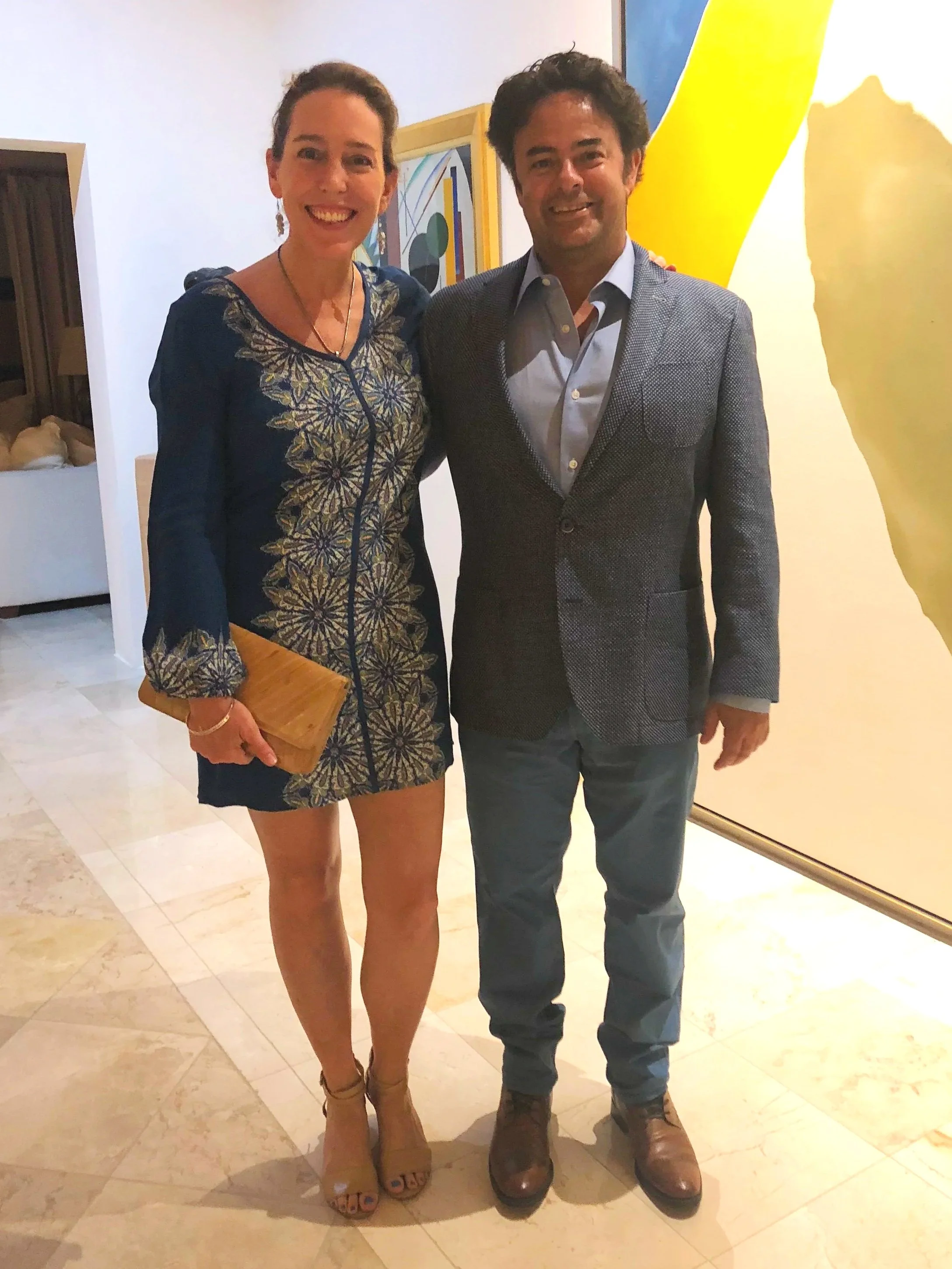 A woman and a man posing together indoors, smiling. The woman wears a patterned dress and heels, holds a clutch purse, and has earrings and a necklace. The man wears a blazer, shirt, jeans, and boots. They stand on a tiled floor with a painting and a patterned wall in the background.