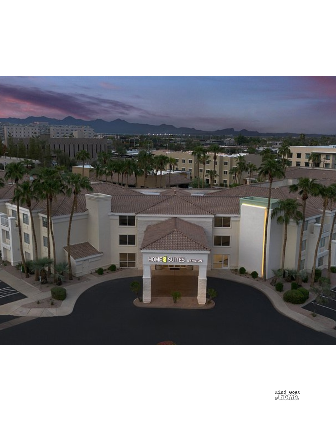 An aerial view of a hotel complex called 'Home2 Suites by Hilton' with palm trees, surrounding low-rise buildings, and mountains in the background during dusk.