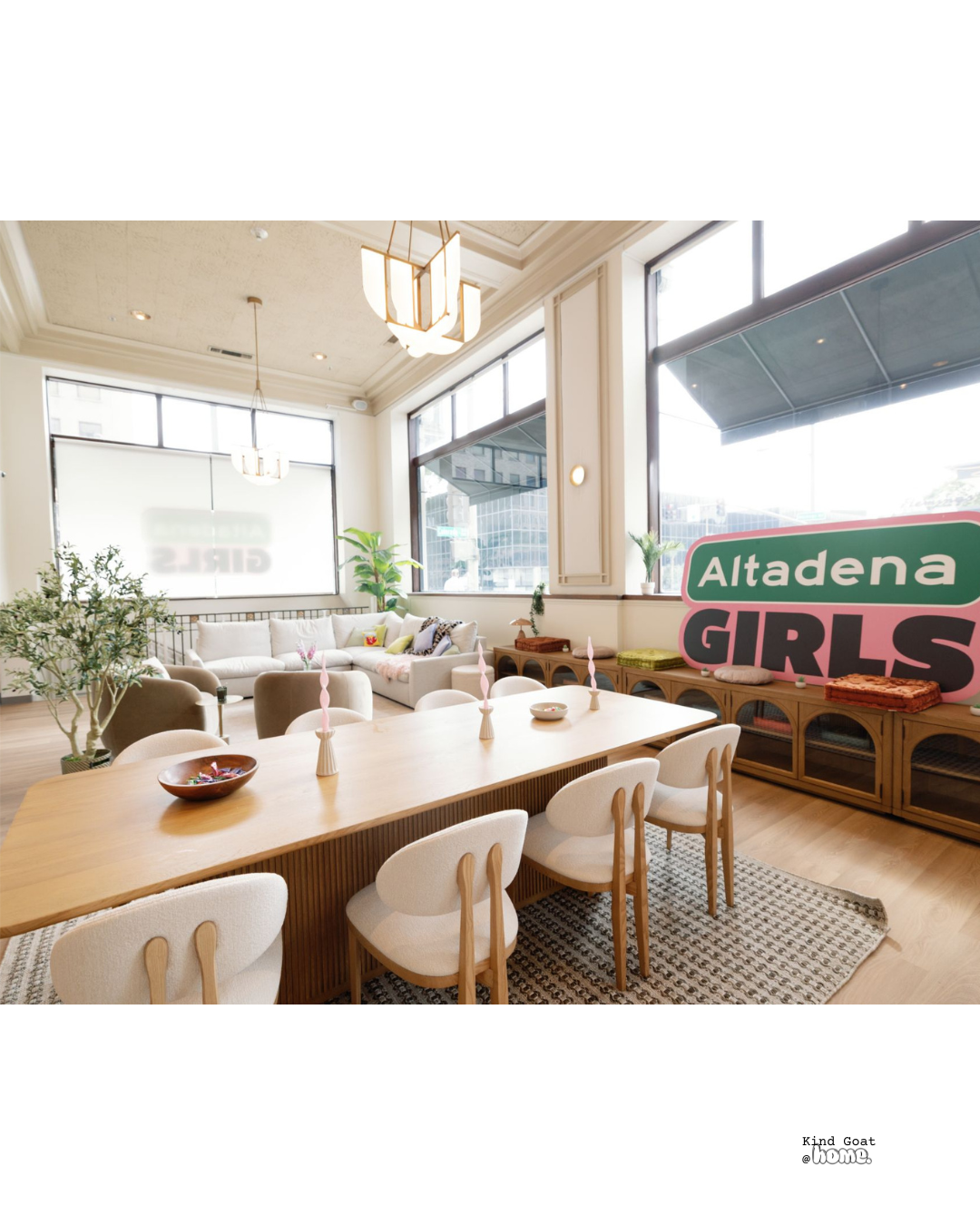 Bright, modern living room with large windows, beige sofa with colorful pillows, wooden coffee table, potted plants, and decorative candles on a dining table. There is a sign that says 'Altadena GIRLS' and some cushions on a wooden sideboard.