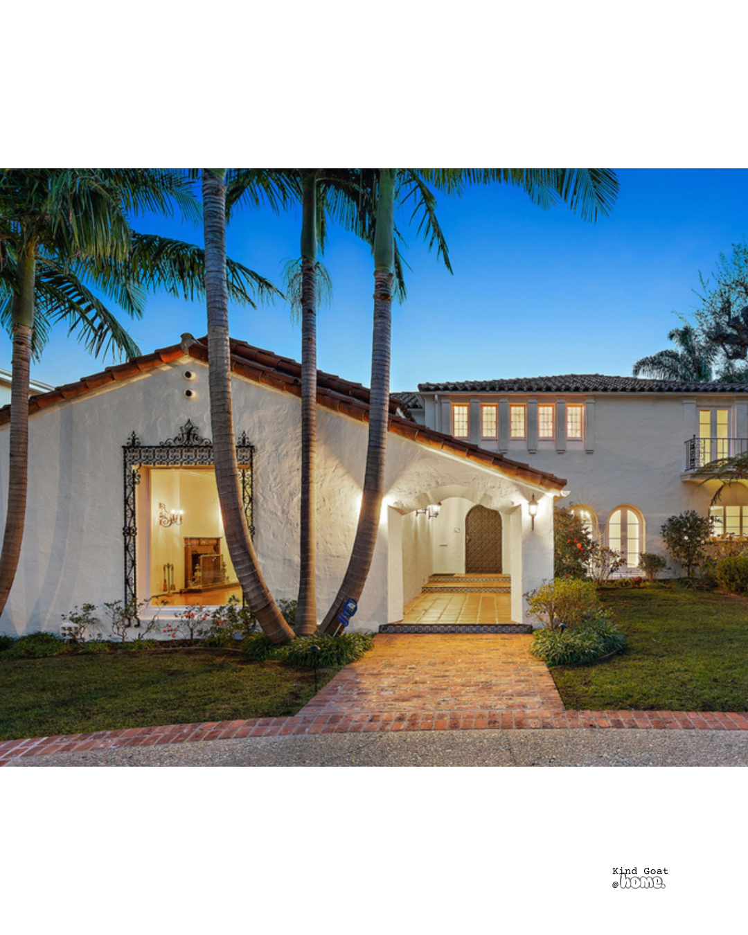 Exterior view of a white Mediterranean-style house with a brick walkway, surrounded by palm trees and greenery, during dusk with lit windows and door.
