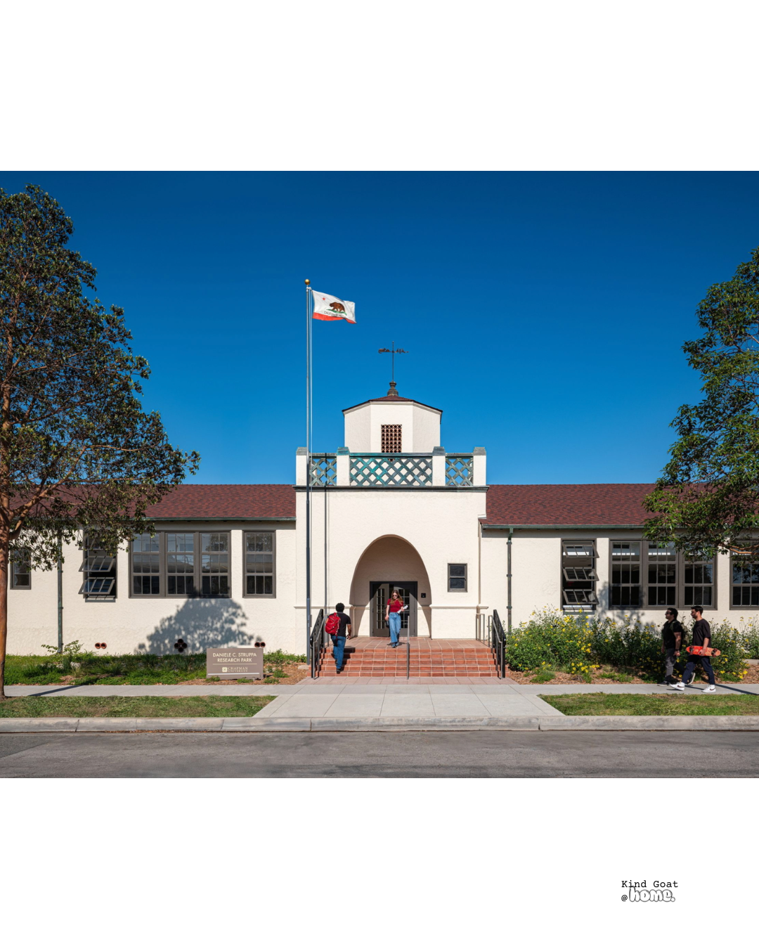 A white building with a red-tiled roof, a small tower with a weather vane, and a flagpole with the California flag. There are three people on the steps and two people walking on the sidewalk in front of the building. The building has sign that reads 'DANIEL C. STRUPPA RESEARCH PARK'.