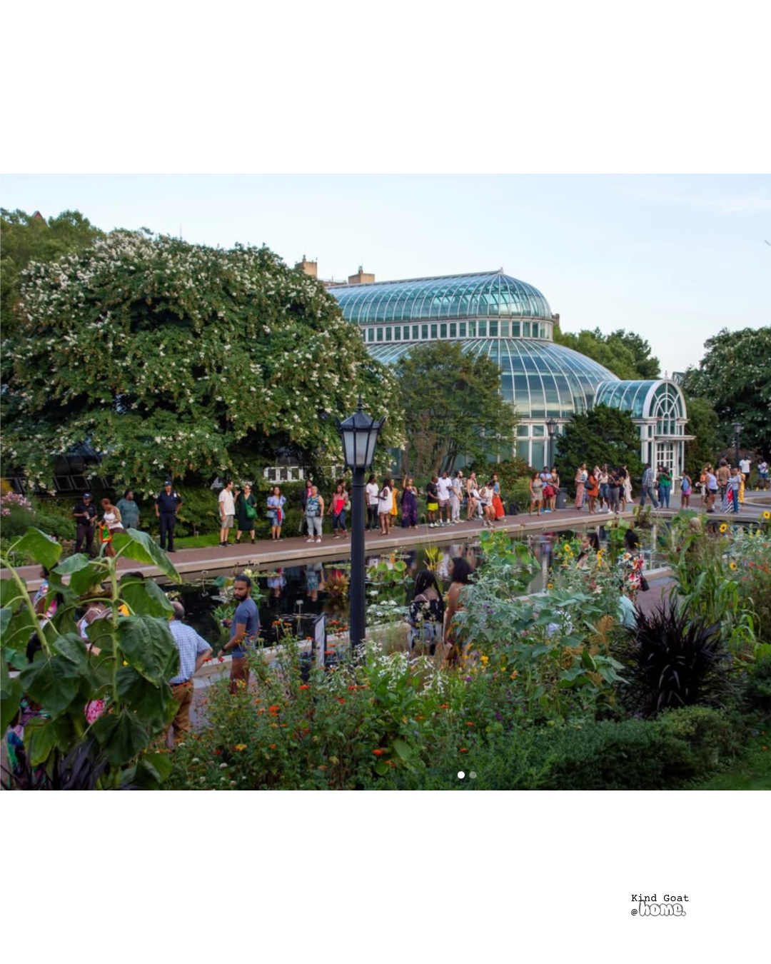 People gather outside a large glass conservatory with lush greenery and blooming trees, near a pond with flowers and plants, under a clear sky.
