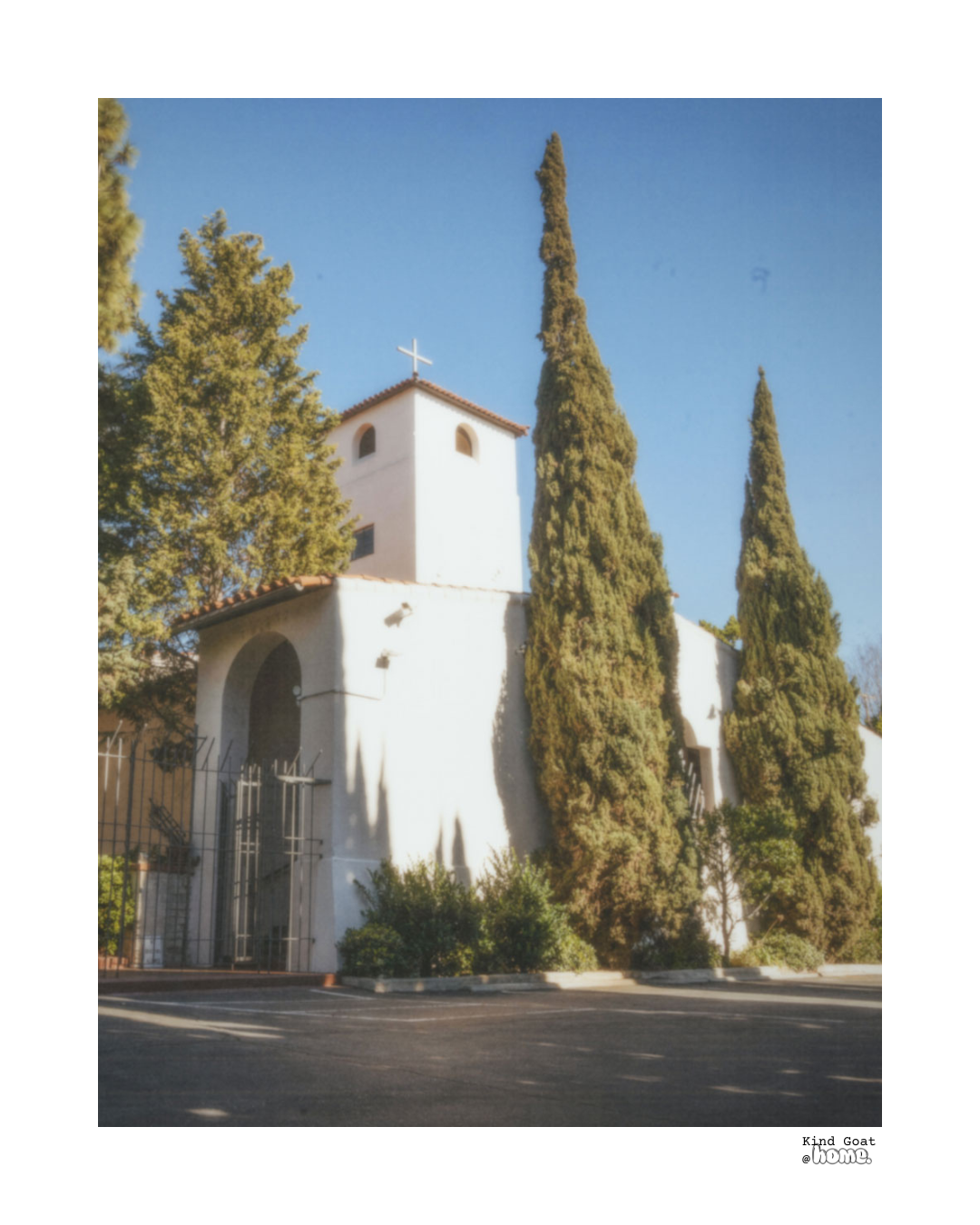 White church with a cross on top, surrounded by tall cypress trees and greenery, under a clear blue sky.