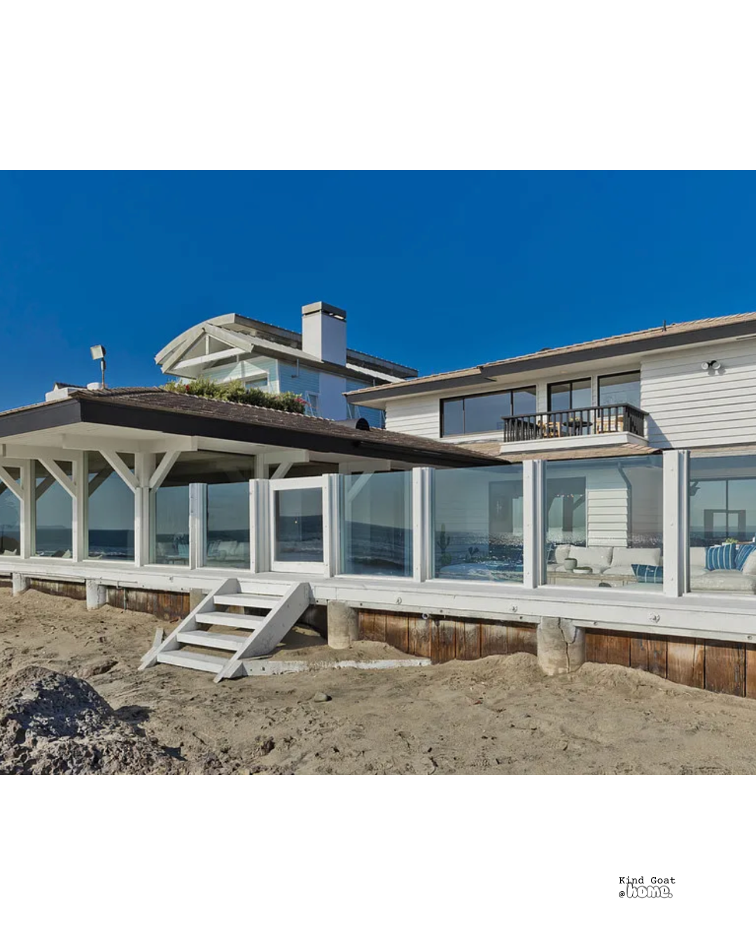 Beach house with white exterior, large windows, and a screened porch area, situated on sandy ground with a clear blue sky overhead.