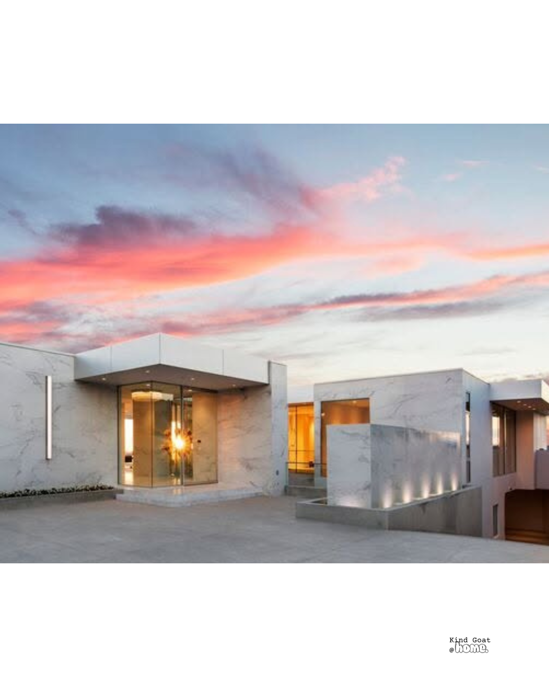 Modern white marble house with large glass windows and a terrace at sunset with pink and orange clouds in the sky.
