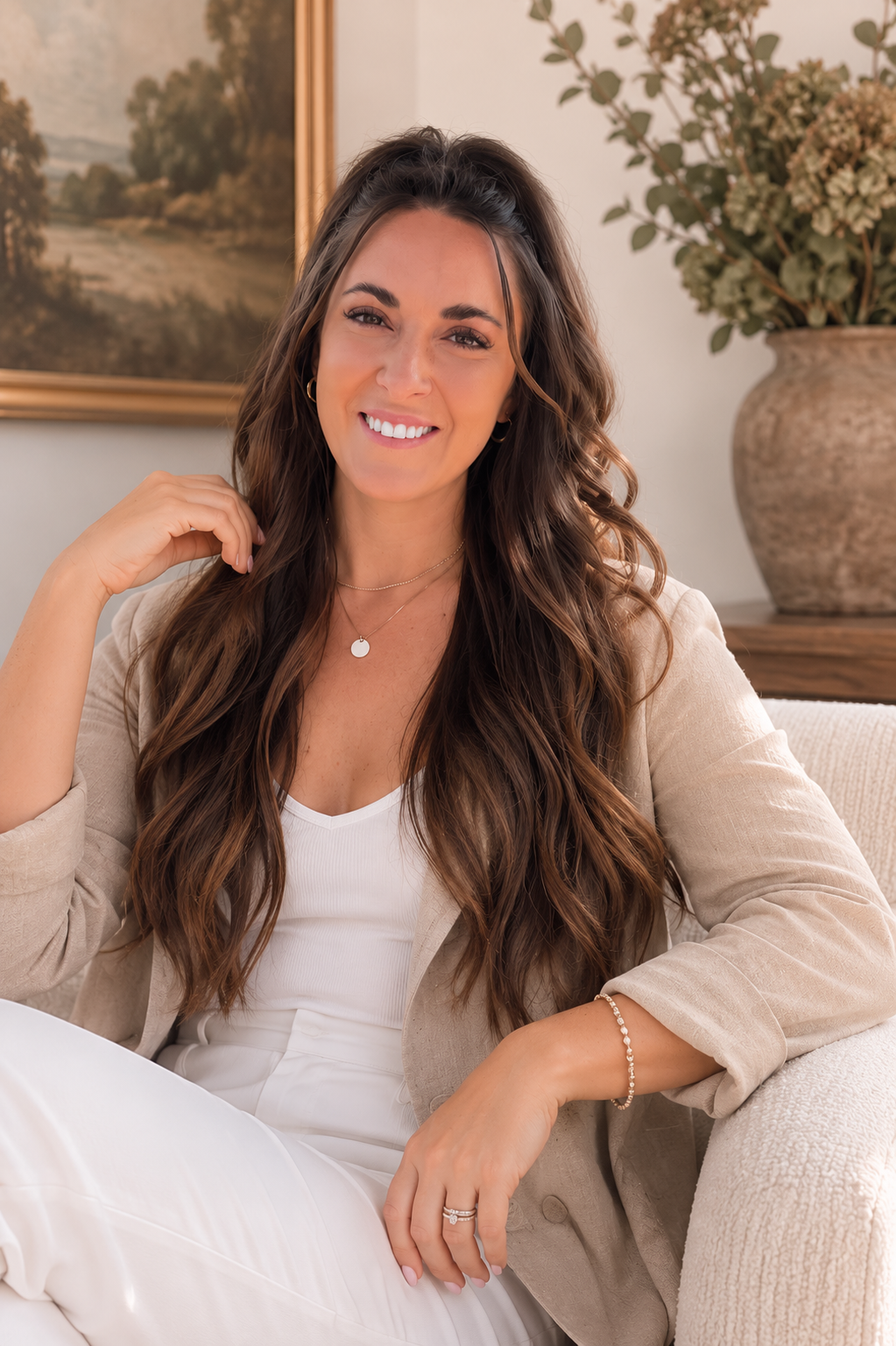 A smiling woman with long, wavy brown hair sitting on a beige couch in a living room, wearing a white top and light-colored blazer, with a large flowerpot and a landscape painting in the background.