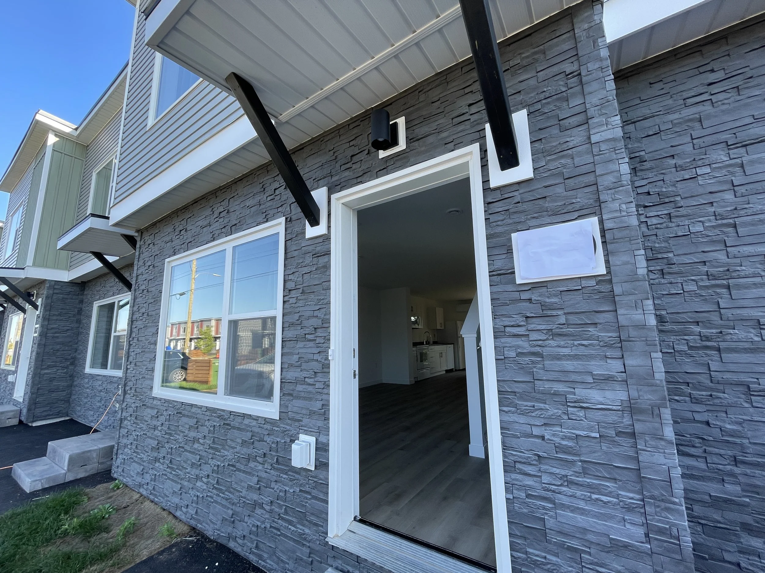 Exterior entrance of a newly constructed townhouse unit at Mount Hope Village in Dartmouth, featuring grey stone veneer siding, an open front door showing the interior wood flooring, and modern outdoor fixtures.