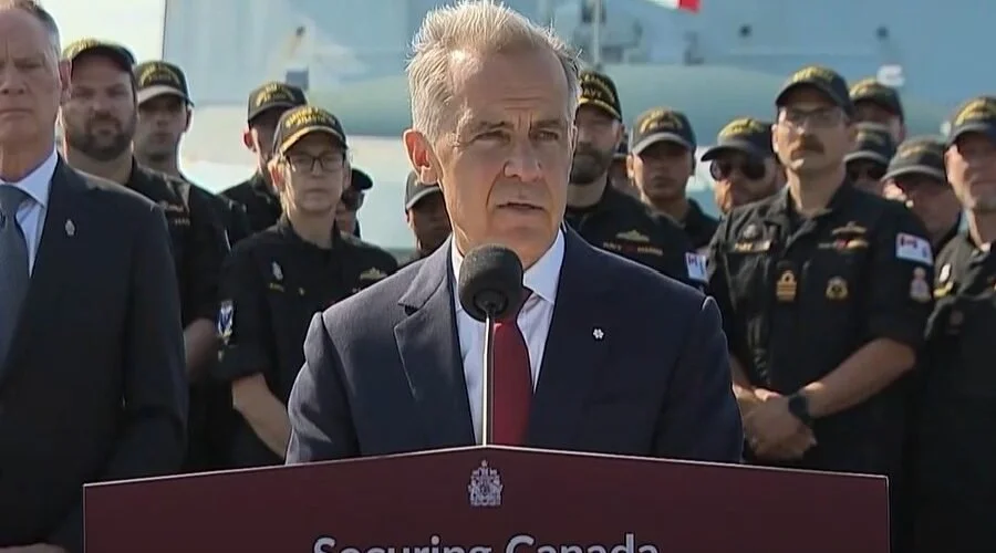 Canadian political leader Mark Carney delivering a speech at a podium branded "Securing Canada" in front of Royal Canadian Navy personnel and a military vessel in Halifax.
