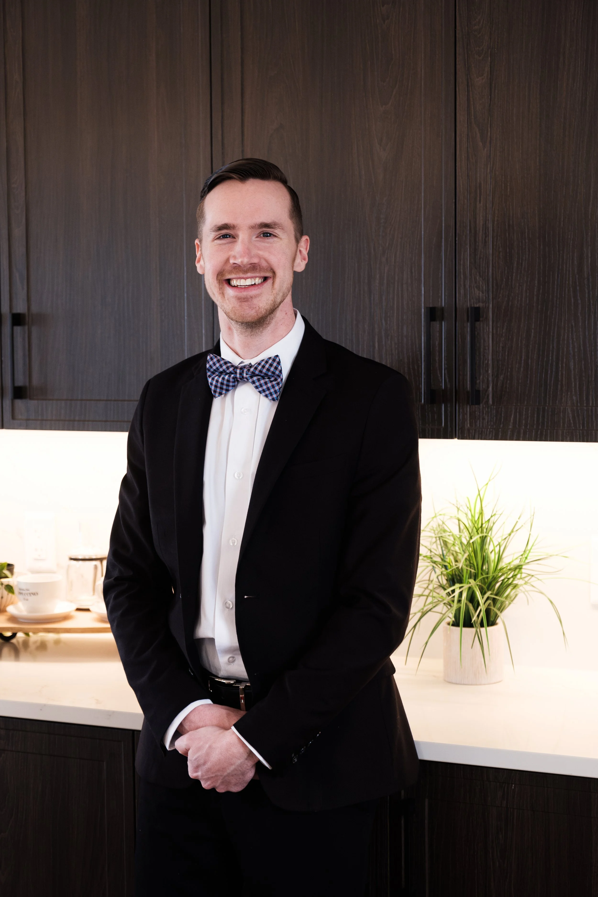 Zach Surette, a local Halifax Real Estate Agent in a black tuxedo with a bow tie, smiling and standing in a kitchen with dark wood cabinets and a white counter, with potted plants and dishes in the background.