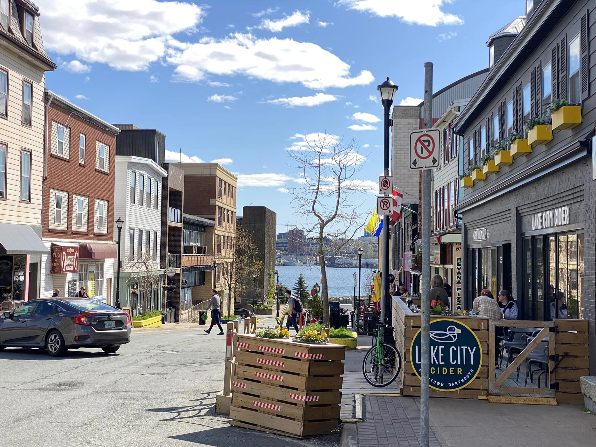 A street view in Lake City with buildings, pedestrians, parked cars, and a view of a body of water in the background on a clear, sunny day.