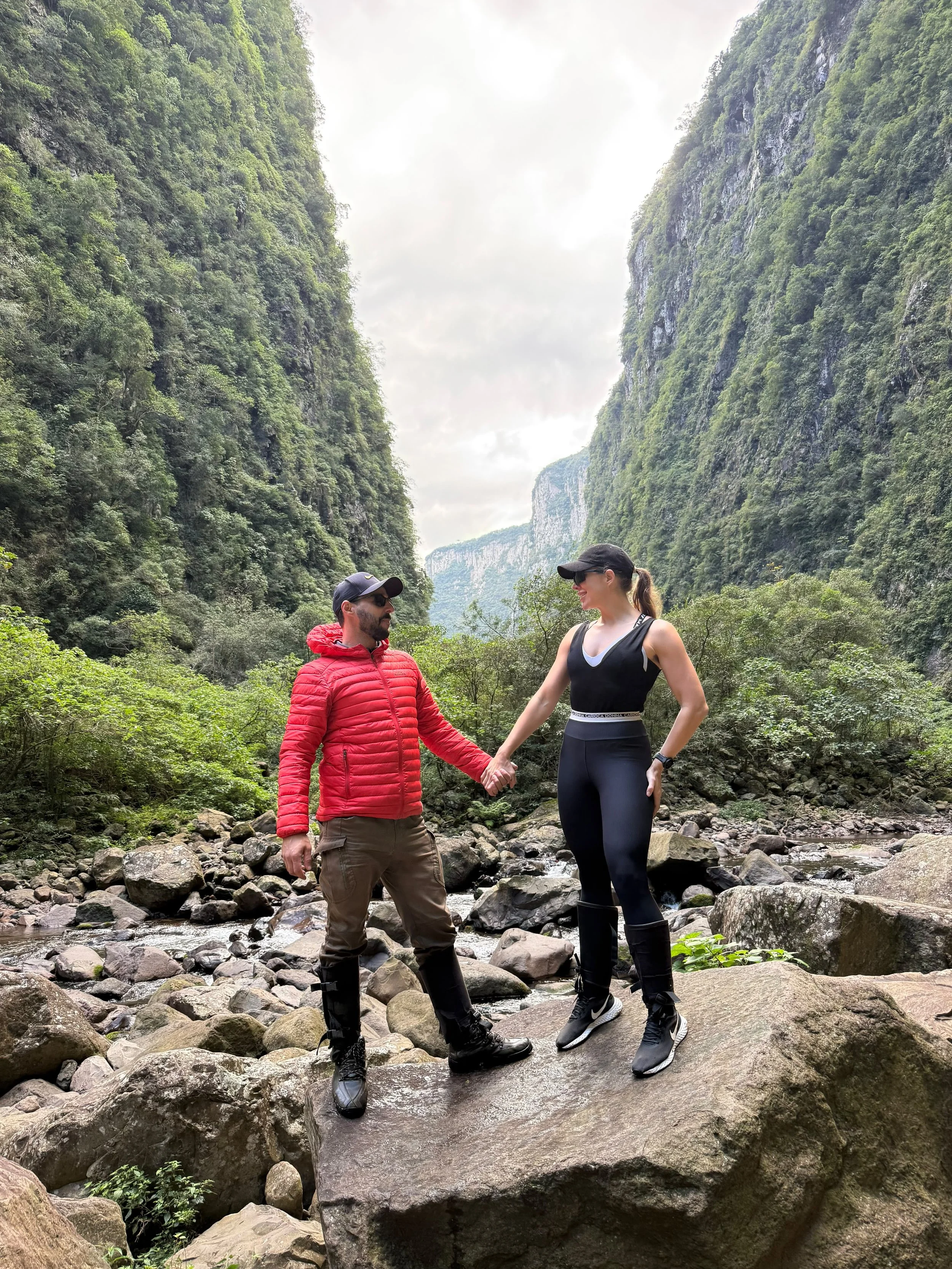 Casal explorando uma cachoeira na floresta, segurando as mãos e olhando um para o outro.