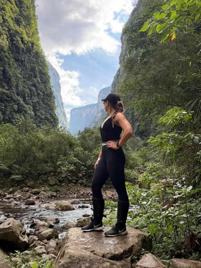 Mulher em roupas esportivas em uma cachoeira rodeada por vegetação densa, com paredões de rocha ao fundo.