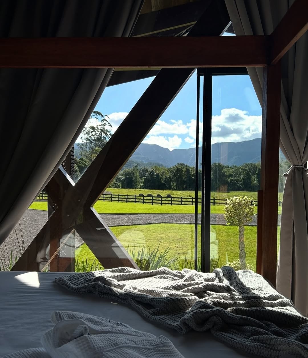 Janela de um quarto com vista para um campo verde, árvore e montanhas ao fundo, com cortinas abertas e uma cama com cobertor e toalha dobrada na frente.